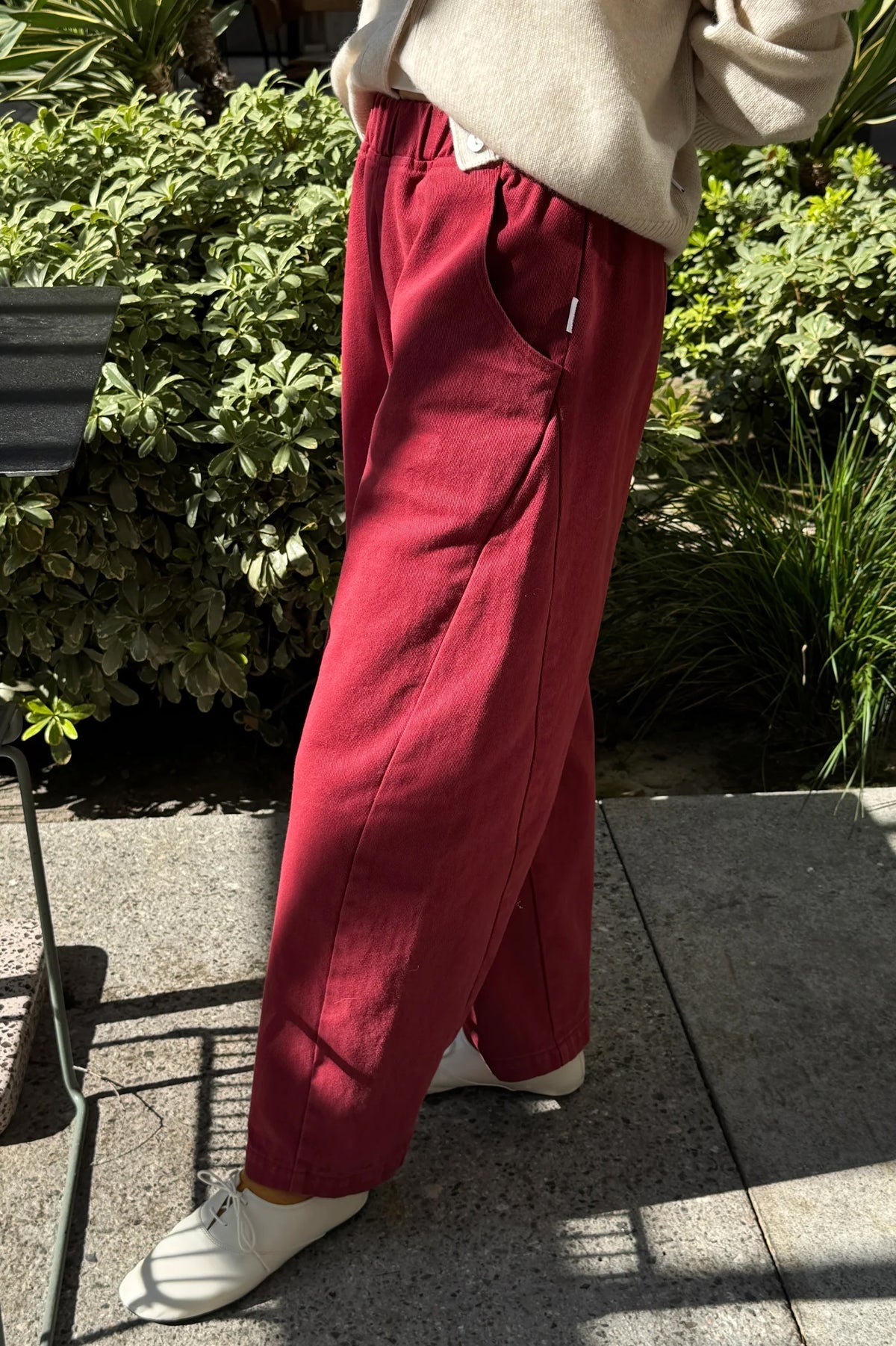 Oxblood Arc Pants, side view showing pocket and curved leg seams, worn with white lace-up shoes