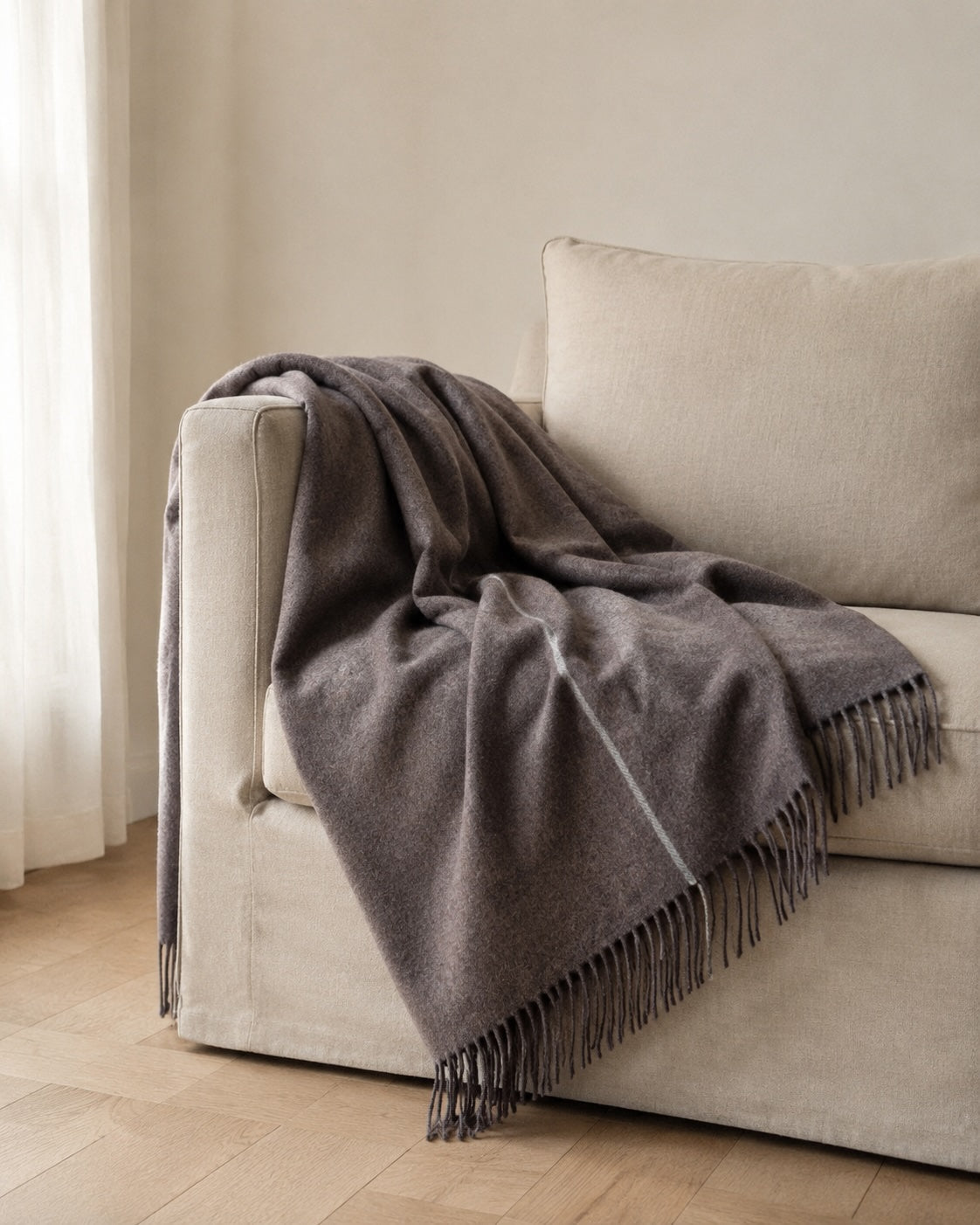 A dark gray wool throw blanket with fringe trim draped over a light beige fabric sofa in a softly lit room.