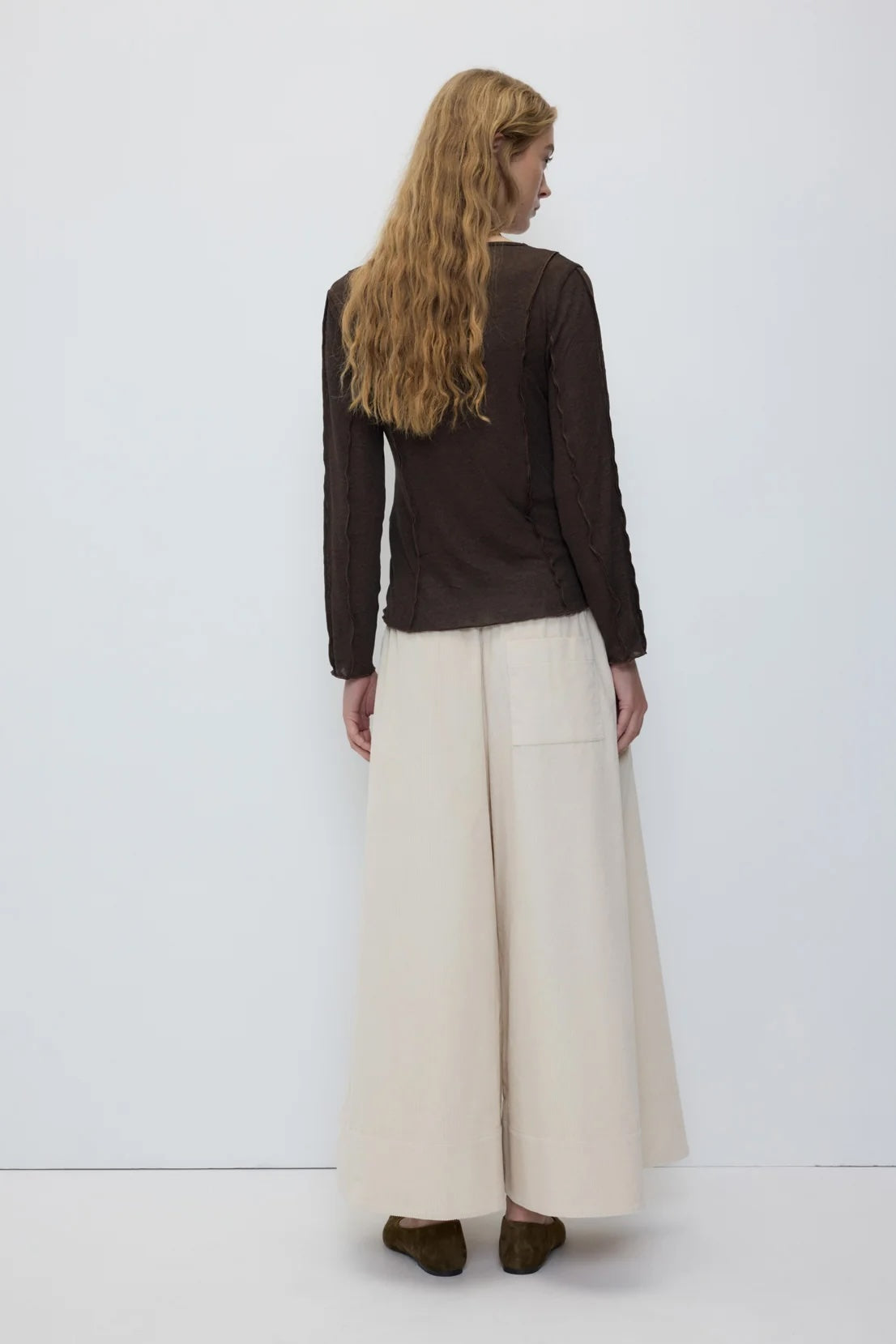 A woman is shown from the back wearing a dark brown long-sleeve top and wide-leg cream trousers, standing against a plain white studio background.