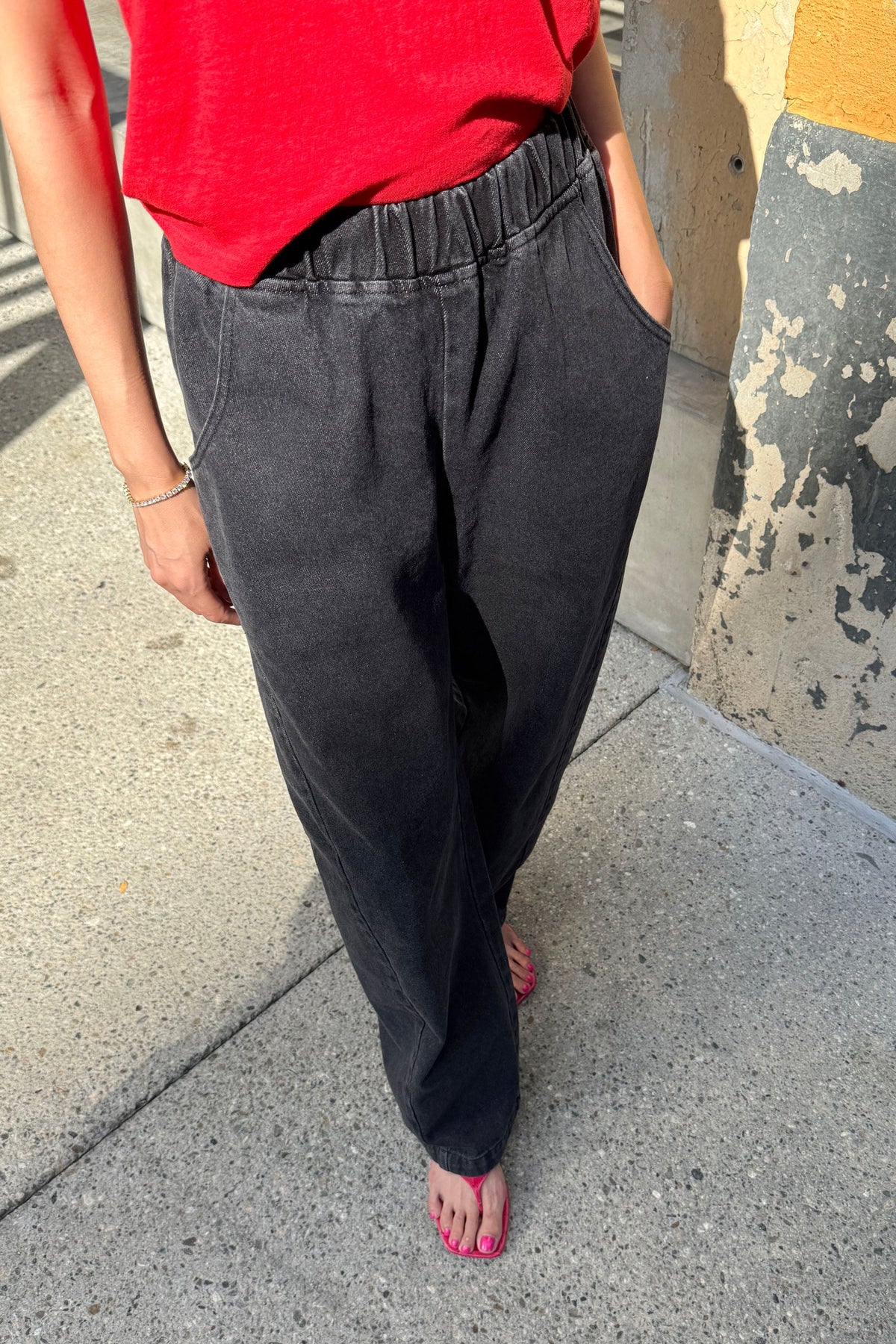 A person wearing loose, dark charcoal gray elastic-waist pants with side pockets, paired with a bright red top and pink sandals, standing on a concrete sidewalk.