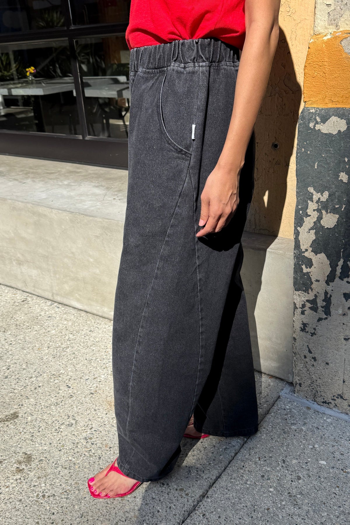 A person wearing wide-leg charcoal gray denim pants with an elastic waistband, standing outdoors on a concrete sidewalk next to a textured wall.