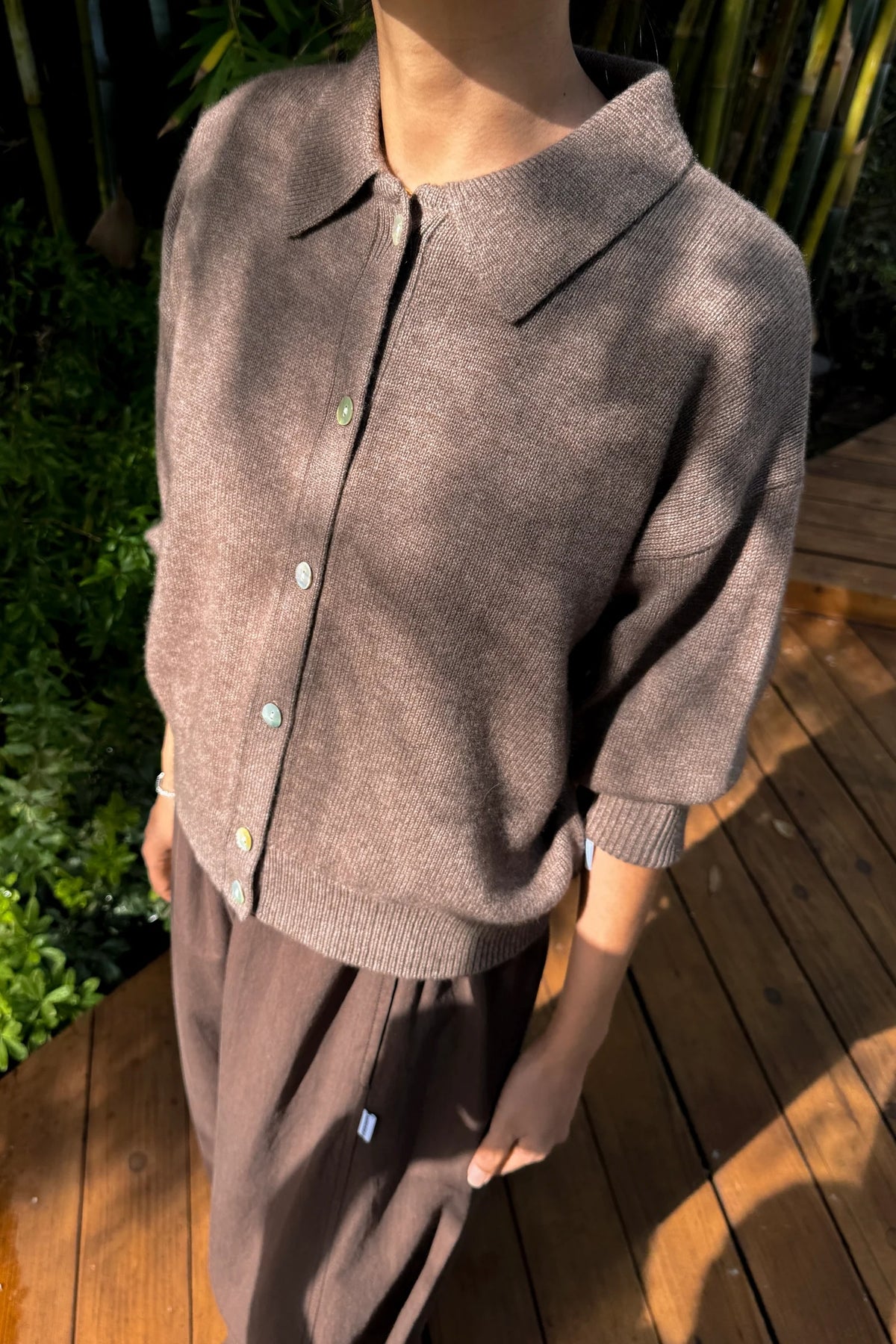 A person wearing a taupe-brown knit cardigan with a pointed collar and three-quarter sleeves, paired with matching brown pants, standing on a wooden deck with green foliage in the background.