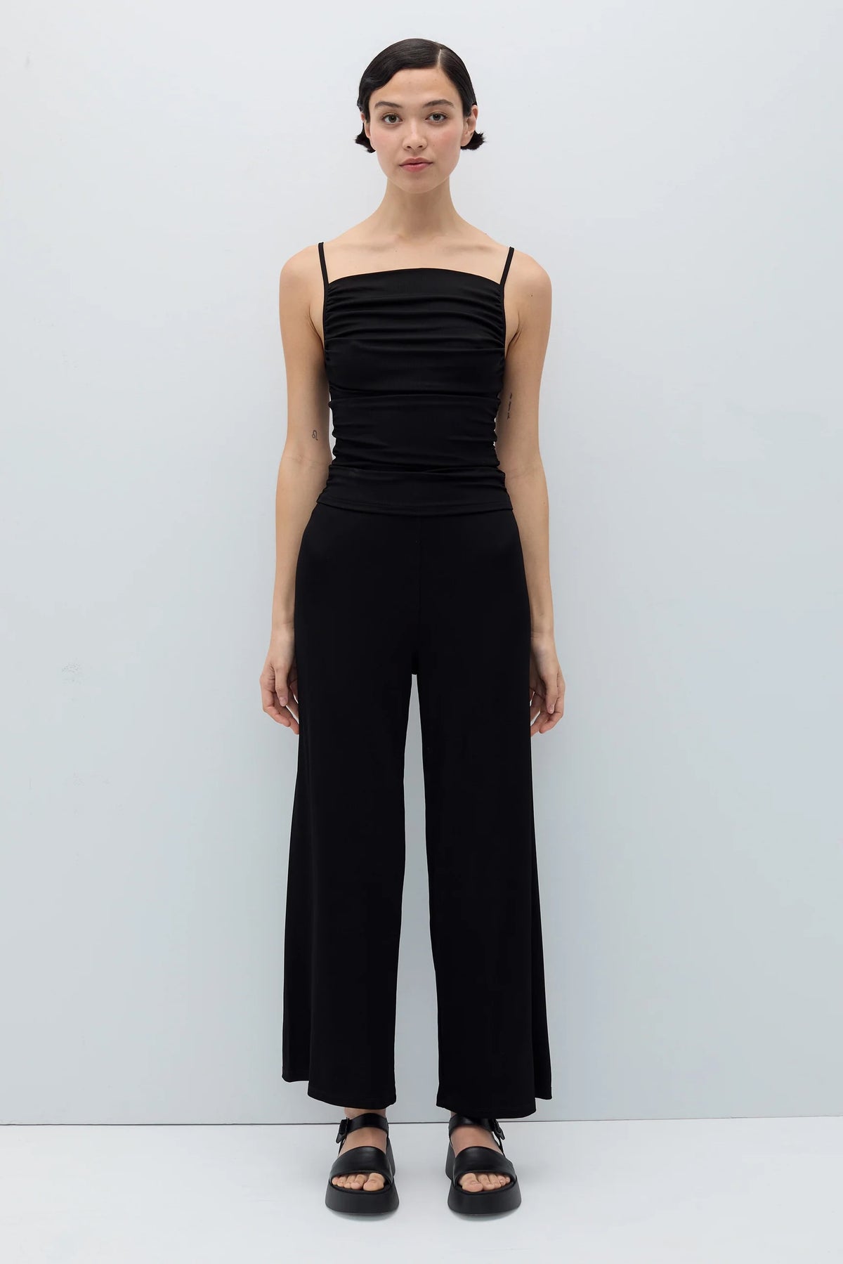 A model stands against a light gray studio background wearing a black sleeveless jumpsuit with thin straps, a ruched bodice, and wide legs, paired with black sandals.