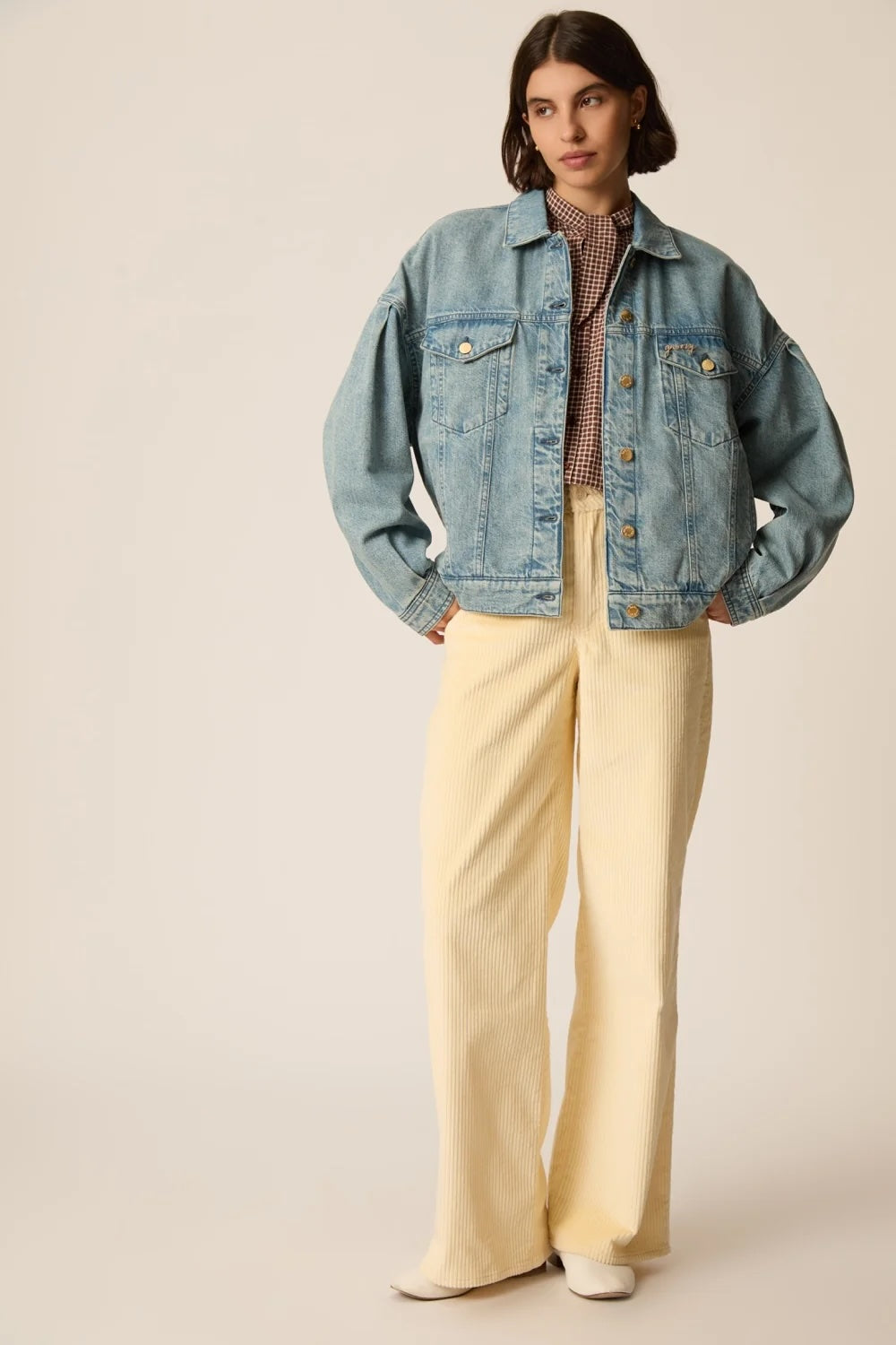 A model stands against a warm beige studio background wearing a light-wash blue denim jacket over a patterned brown shirt, paired with cream wide-leg trousers and white shoes.
