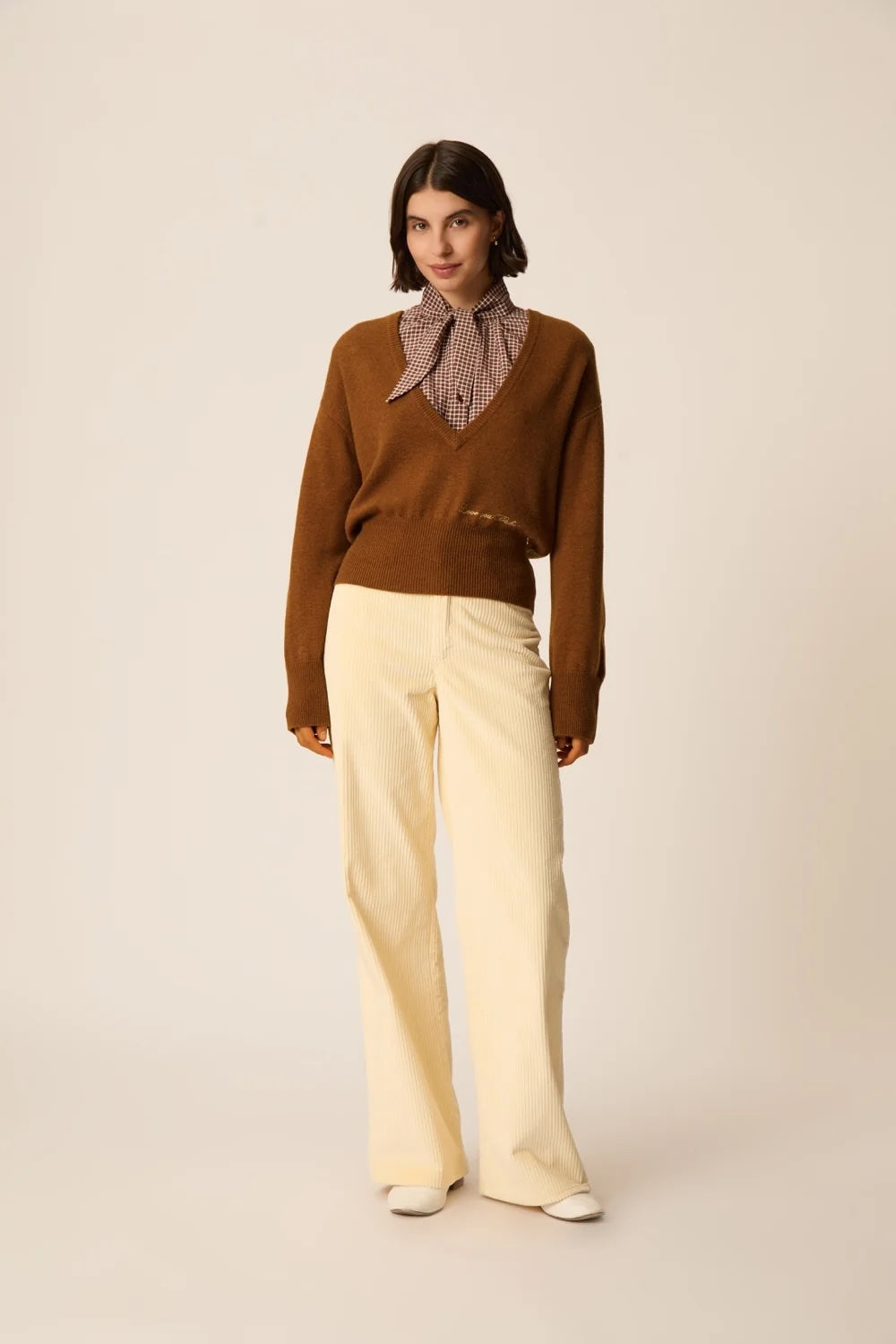A model stands against a plain beige studio background wearing a brown knit sweater over a patterned collared shirt, paired with cream wide-leg trousers and white shoes.