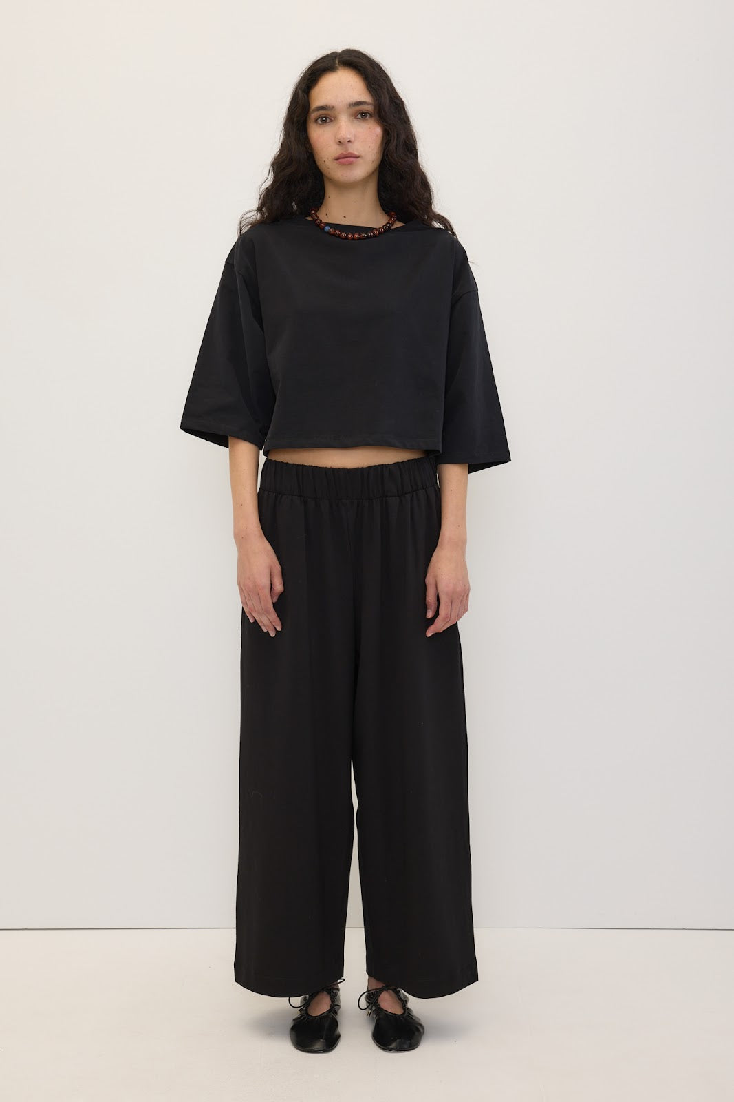A model stands against a plain white studio background wearing a black cropped short-sleeve top with a beaded neckline and black wide-leg pants.