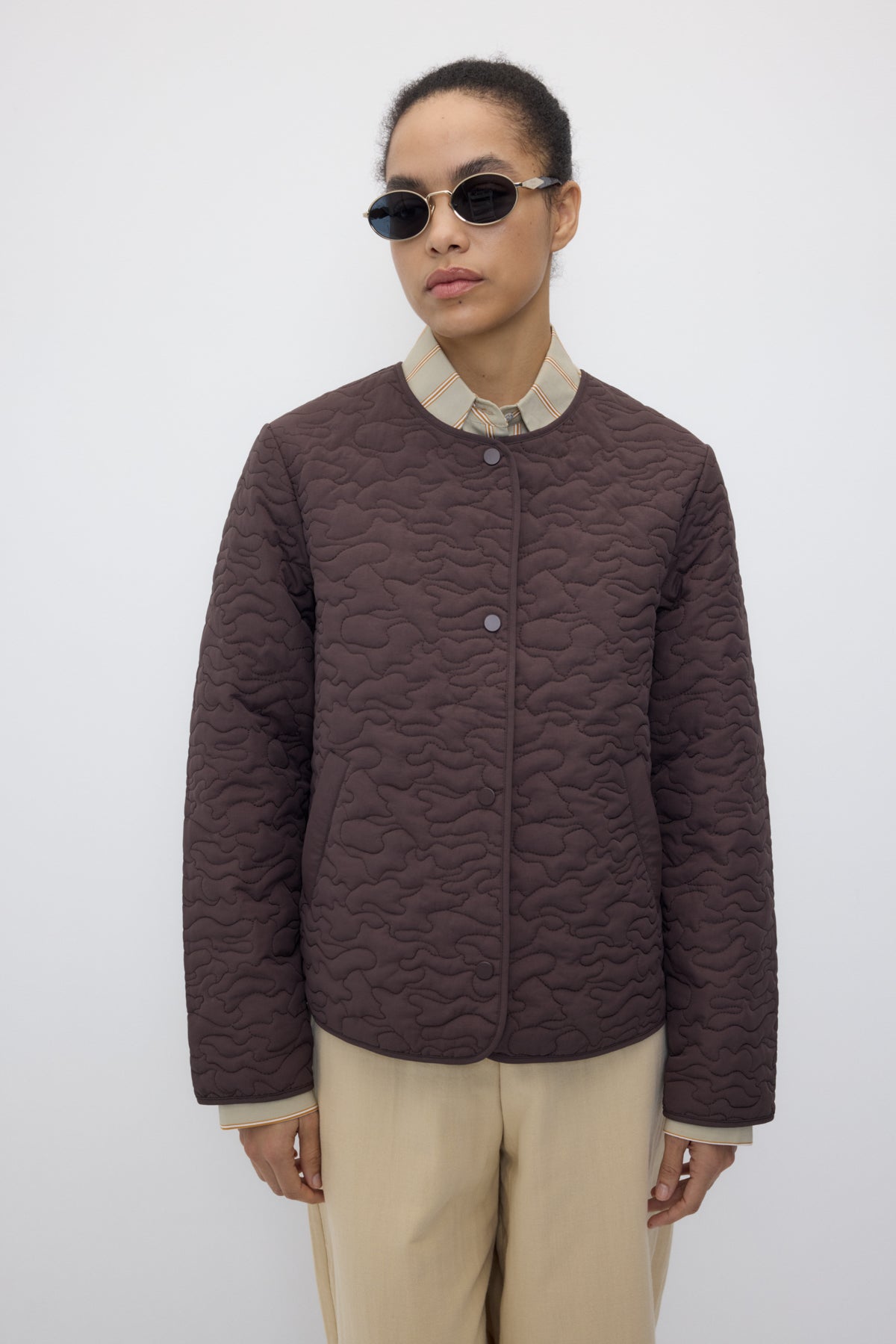 A model wearing a dark brown quilted jacket with a beige collar, paired with beige trousers and round sunglasses, against a plain light gray background.