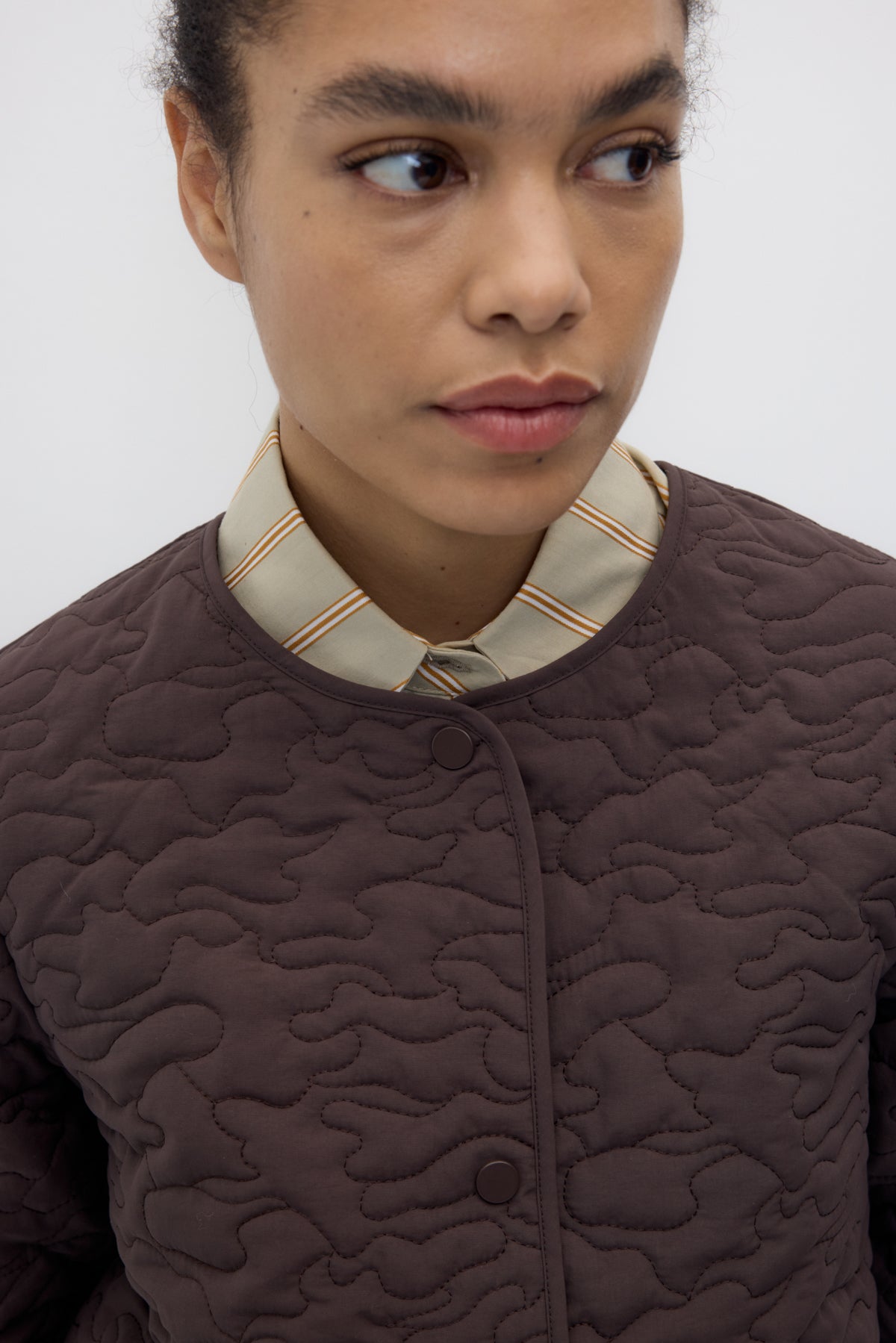 Close-up of a model wearing a dark brown quilted jacket with a patterned beige and brown striped collar over a matching striped shirt.