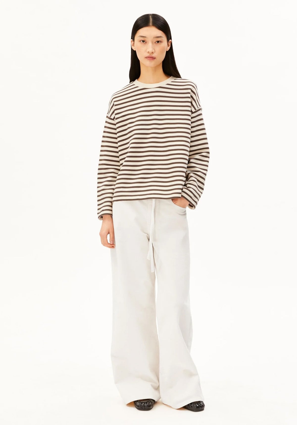 A model stands against a plain white background wearing a brown and white horizontally striped long-sleeve knit top with white wide-leg pants.