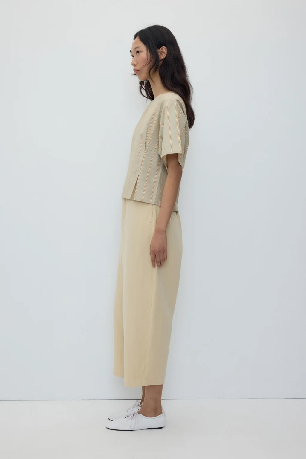 A woman stands in profile wearing a light beige two-piece outfit with a short-sleeve cropped top and matching wide-leg cropped pants, paired with white sneakers against a plain white studio background.