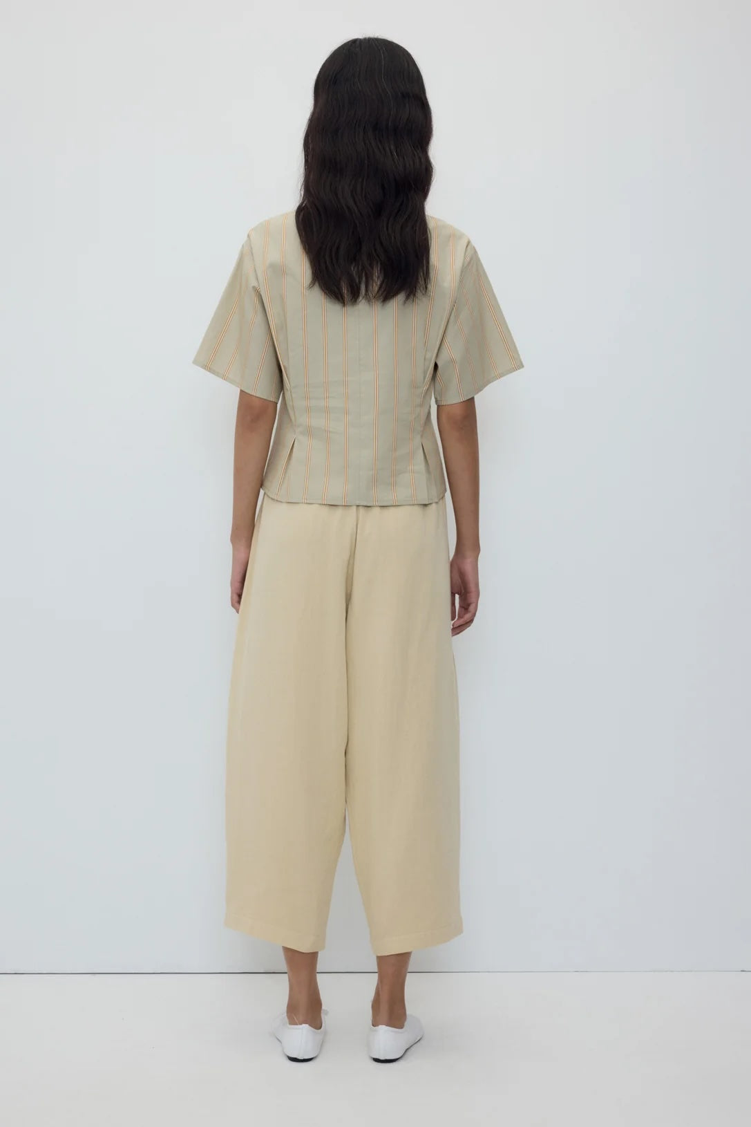 A model viewed from the back wearing a light beige short-sleeve top with thin vertical stripes and matching wide-leg cropped pants, paired with white shoes, against a plain white studio background.