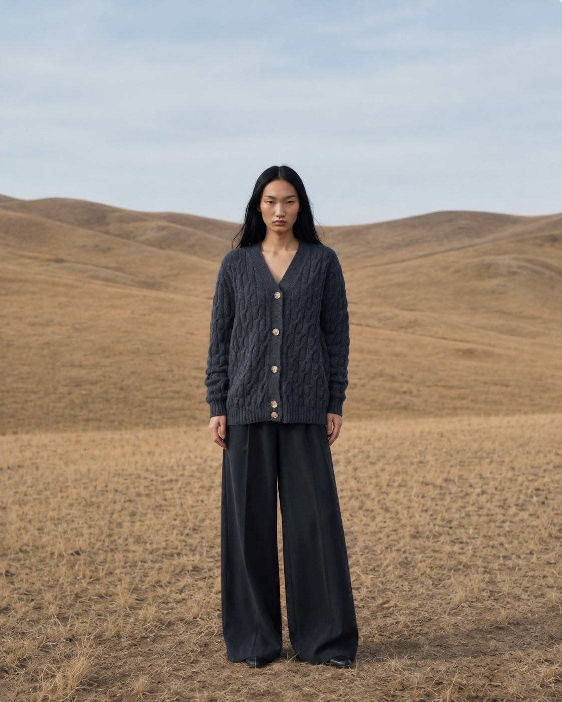 A model stands in a dry, rolling grassy field under a pale sky, wearing a dark gray cable-knit button-front cardigan and matching dark wide-leg trousers.
