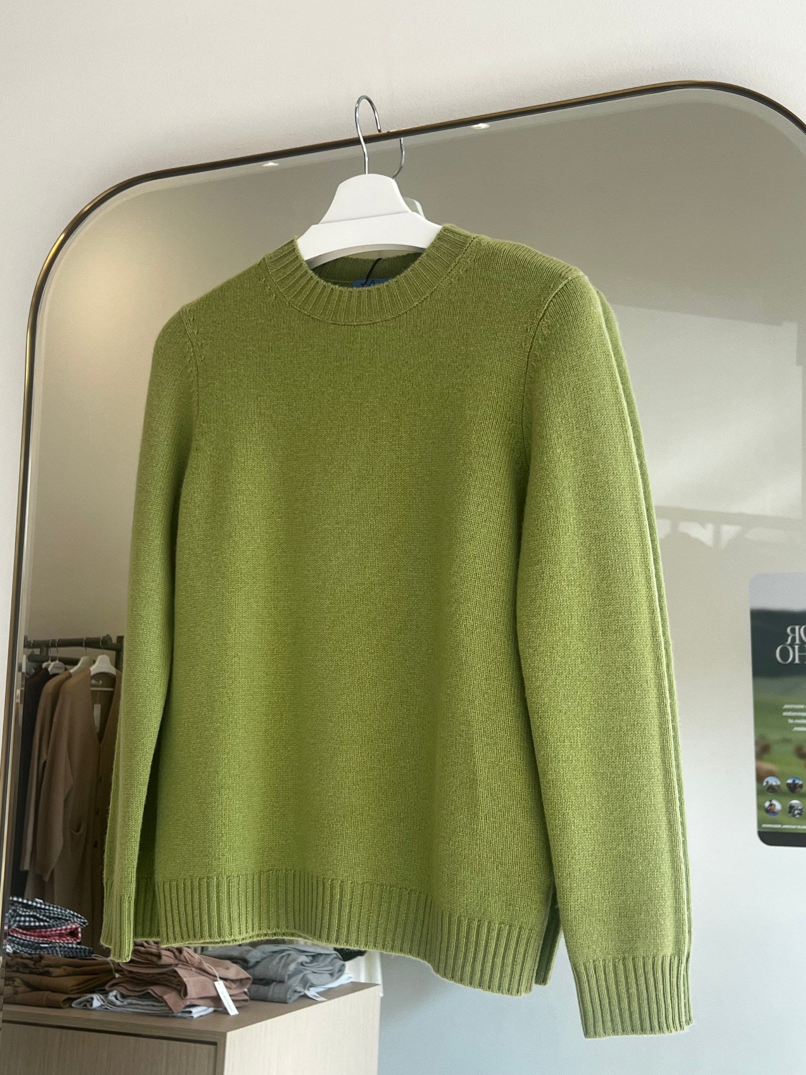 Green round neck cashmere sweater hanging on white hanger in front of mirror in store interior