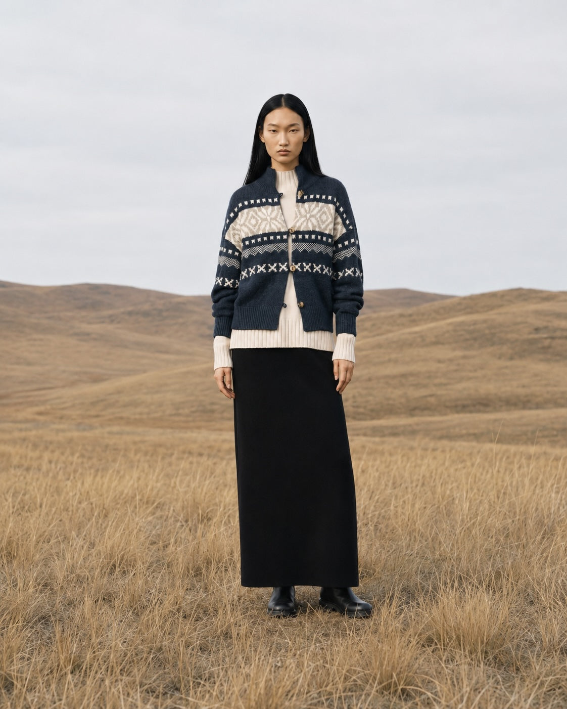 A model stands in a dry grassy field under an overcast sky, wearing a navy and cream patterned knit cardigan over a cream top and a long black skirt.