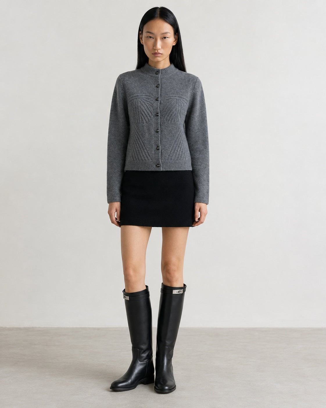 A model stands against a light gray studio background wearing a gray textured knit cardigan with a stand collar and front buttons, paired with a black mini skirt and tall black rain boots.