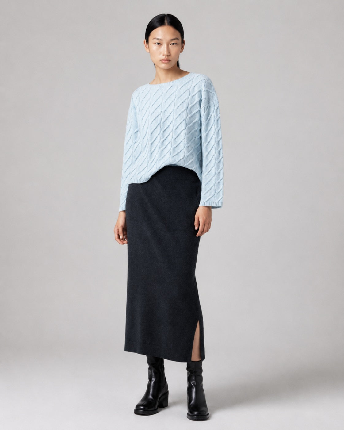 A model stands against a light gray studio background wearing a light blue cable-knit sweater and a black midi skirt with a side slit, paired with black ankle boots.