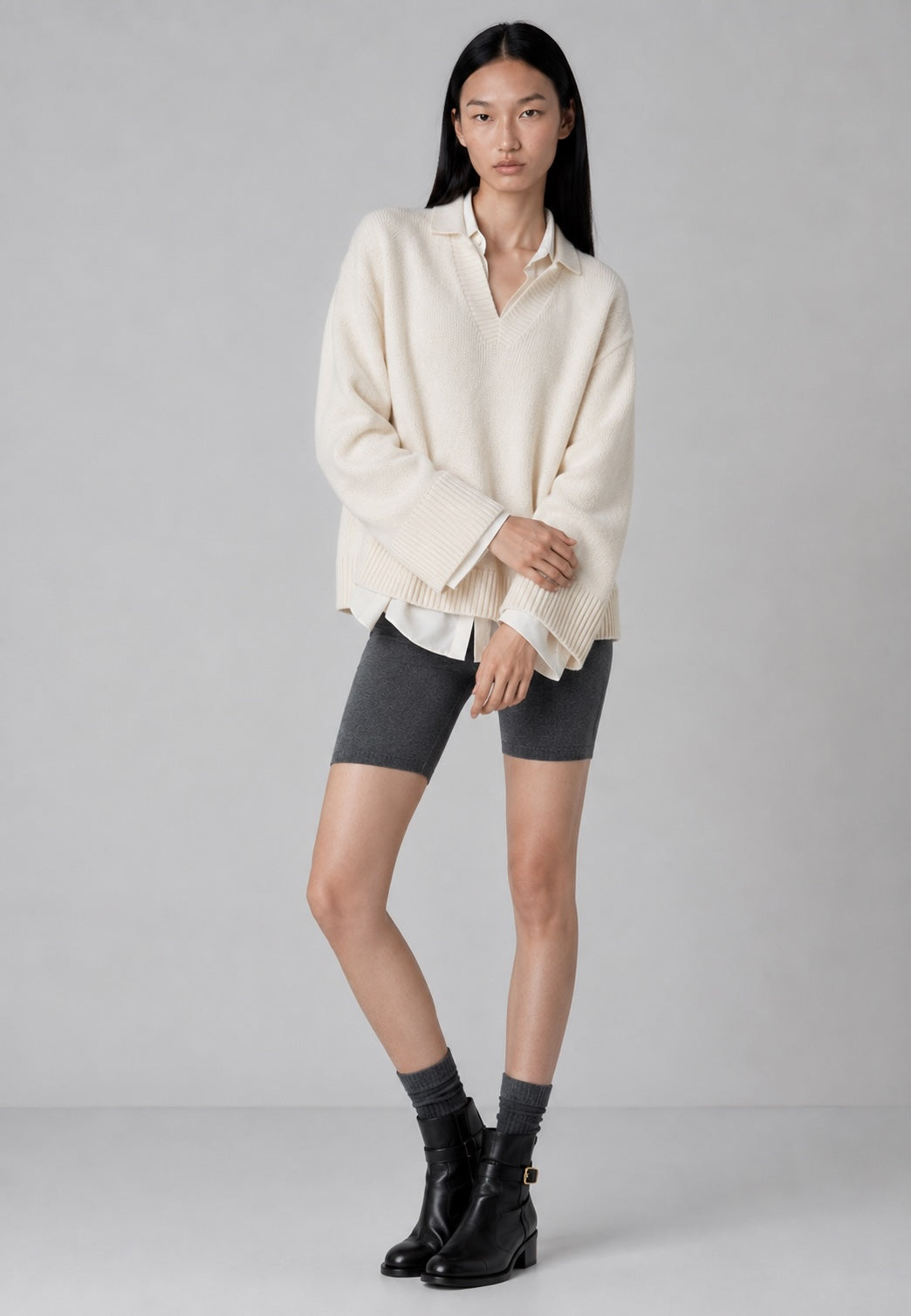 A model stands against a light gray studio background wearing an oversized cream ribbed knit polo sweater, dark gray fitted shorts, dark gray socks, and black ankle boots.