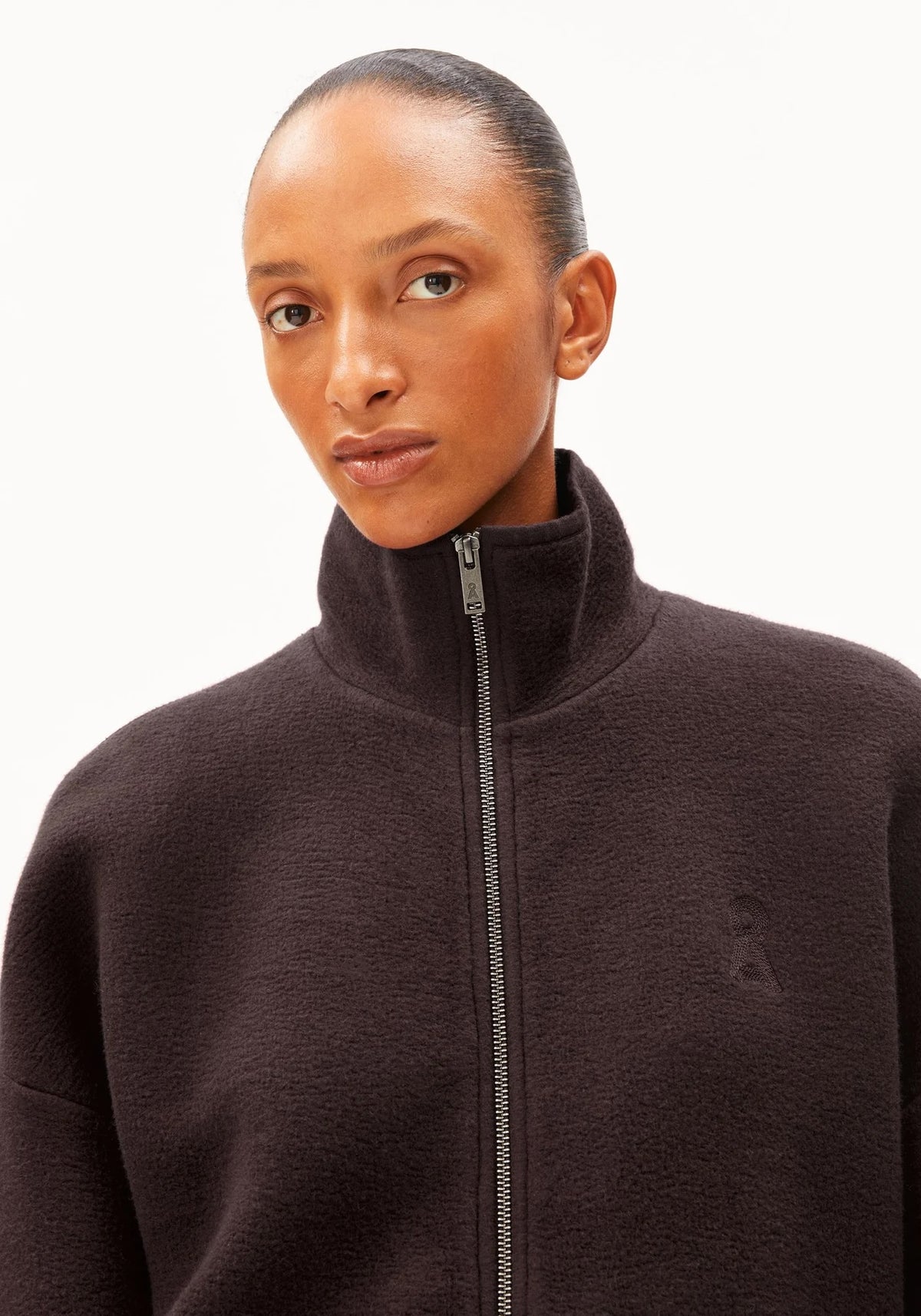A close-up of a model wearing a dark brown high-collar zip-up fleece jacket against a plain white background.