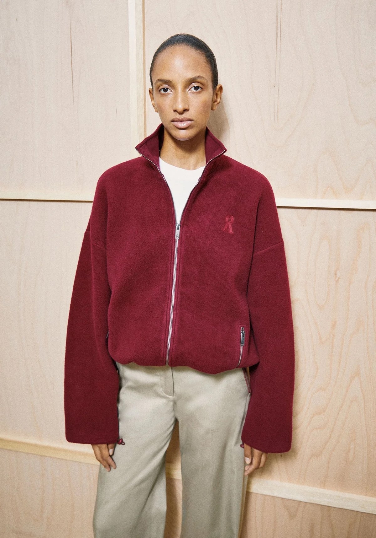 A model wearing a deep burgundy fleece zip-up jacket with a small embroidered logo on the left chest, paired with light beige trousers and a white t-shirt, standing against a light wood-paneled wall.