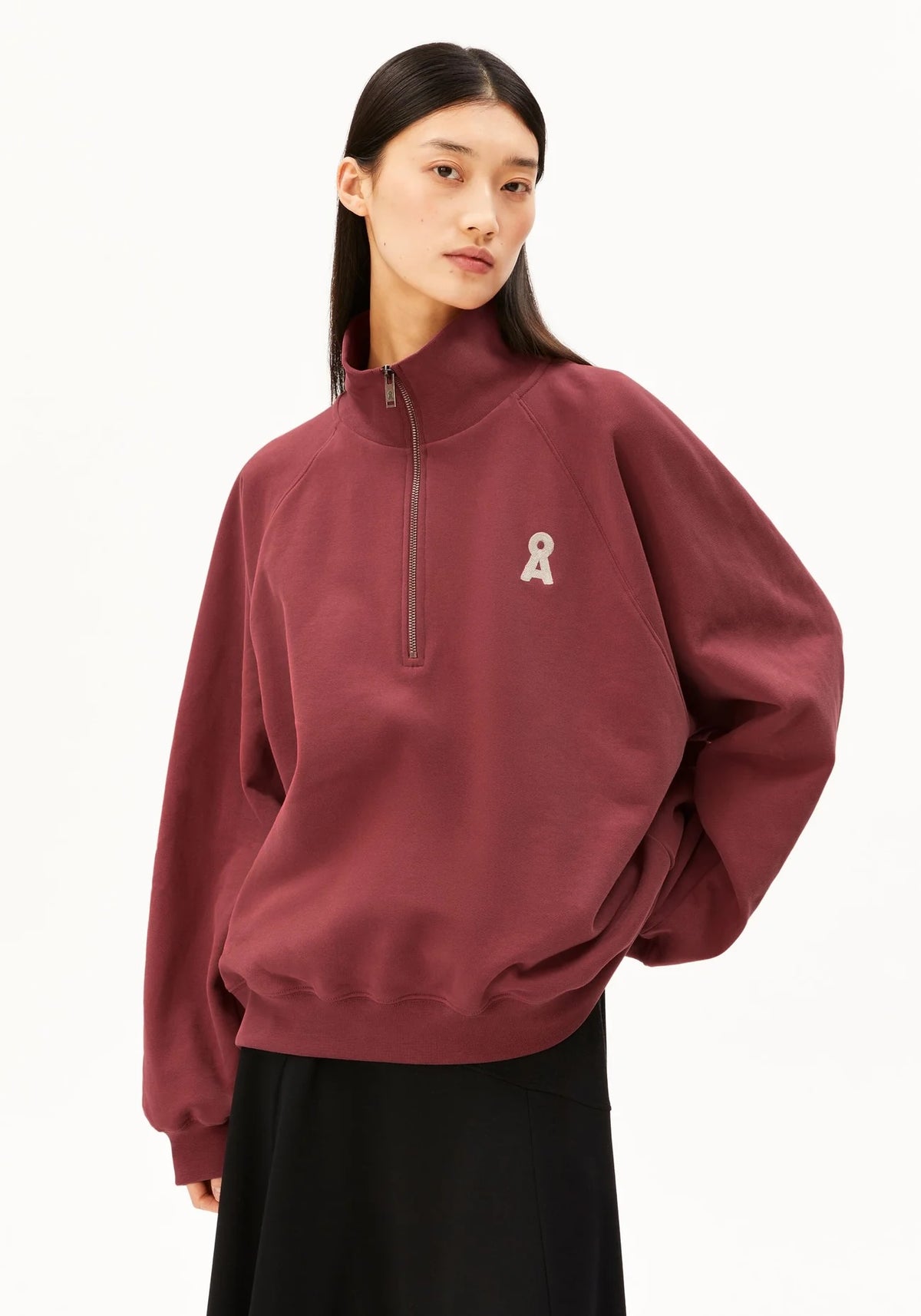 A model wearing a maroon half-zip sweatshirt with a small white ribbon graphic on the left chest, paired with a black skirt, against a plain white background.