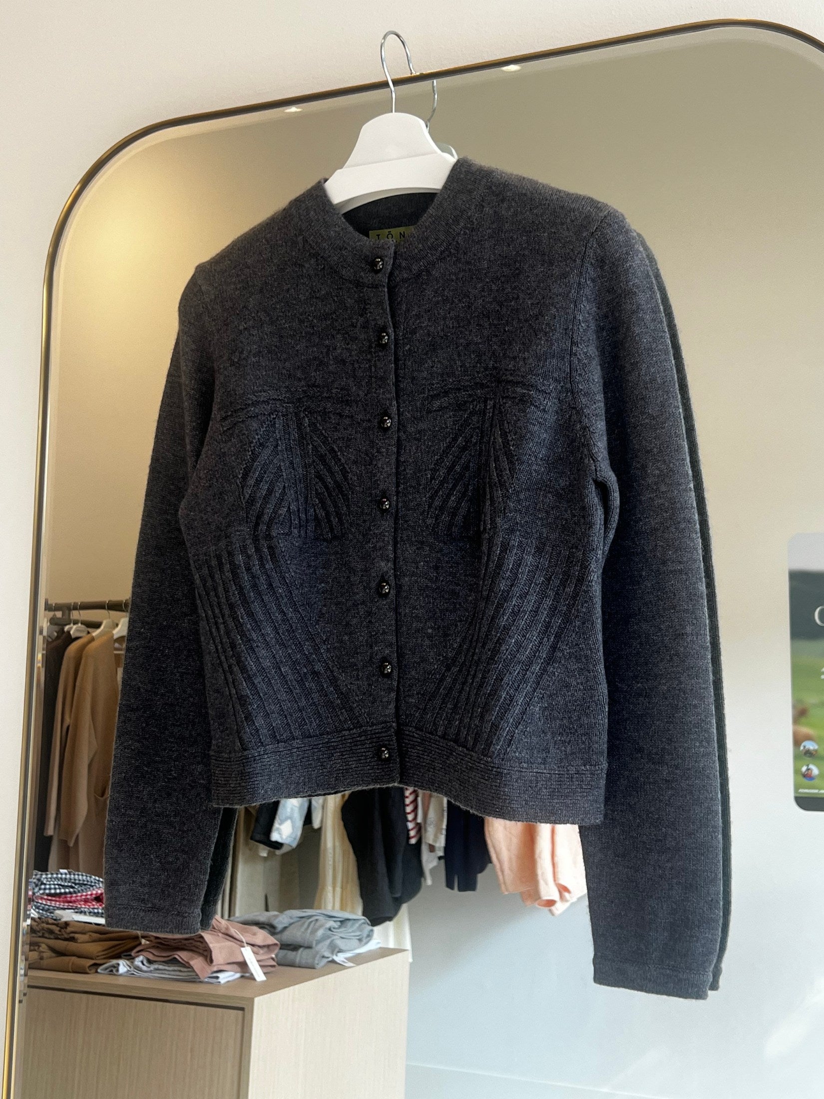Dark gray shift stitch crop button cashmere cardigan hanging on white hanger in a boutique setting