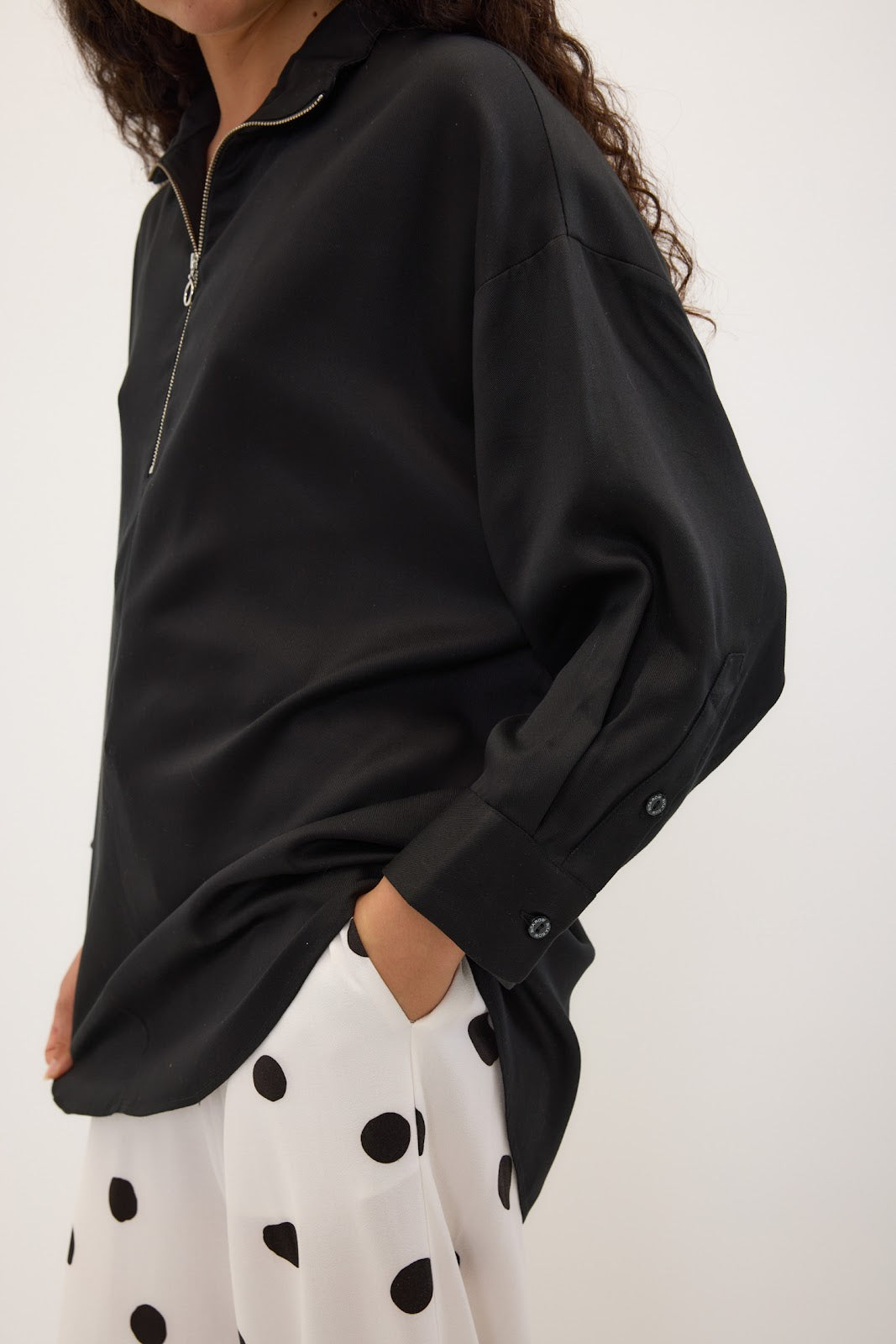 A close-up of a person wearing a black long-sleeve blouse with a half-zip neckline and white pants with black polka dots, against a plain light background.