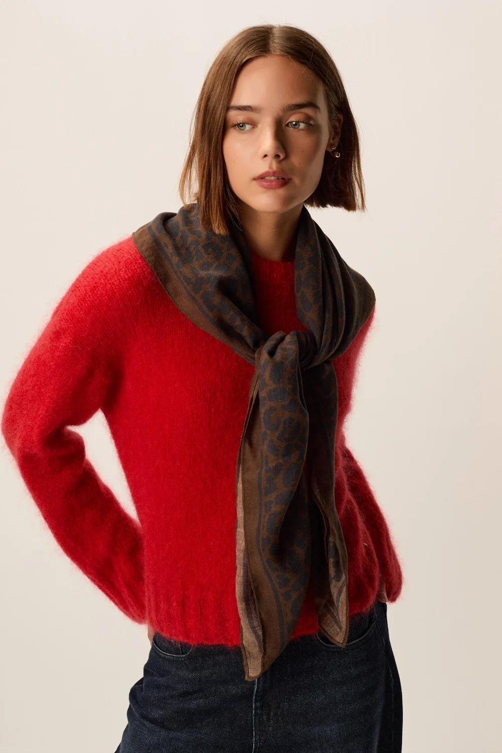 A woman wearing a bright red fuzzy knit sweater and a dark brown patterned scarf, photographed against a plain light beige studio background.