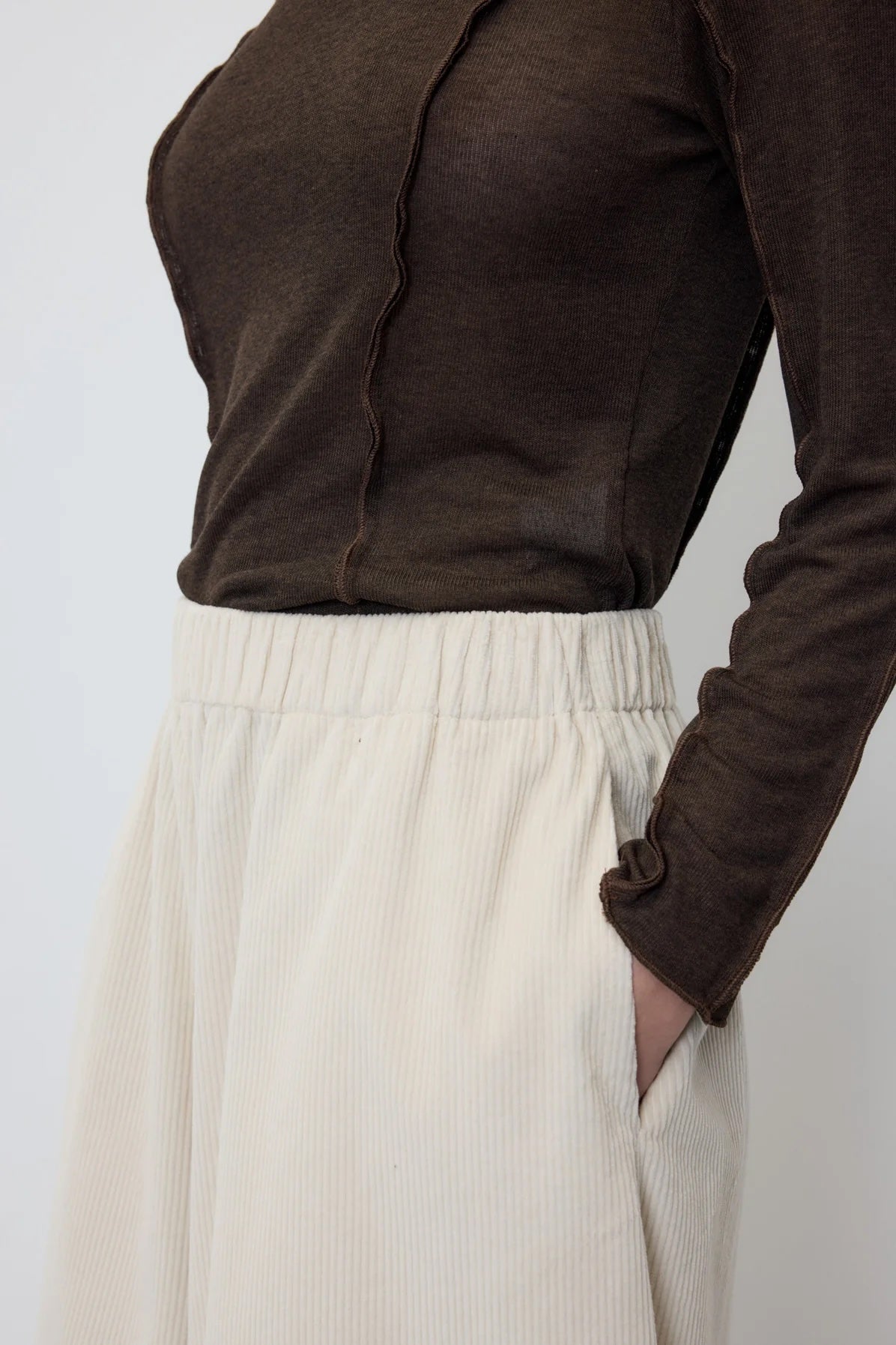 Close-up of a person wearing a dark brown long-sleeve top and cream-colored pleated trousers, with one hand in a pocket.
