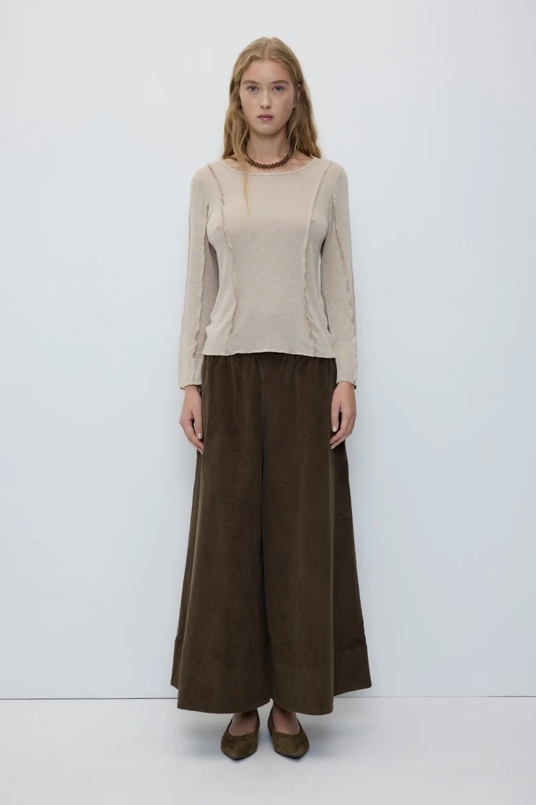 A woman stands against a plain light gray studio background wearing a beige long-sleeve knit top and wide-leg dark brown trousers, with brown pointed flats.
