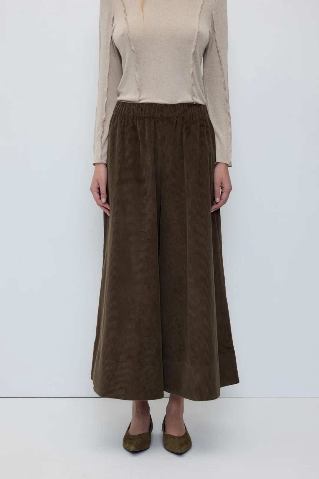 A model wearing a pair of wide-leg brown corduroy trousers, paired with a beige long-sleeve top and olive green shoes, standing against a plain white studio background.