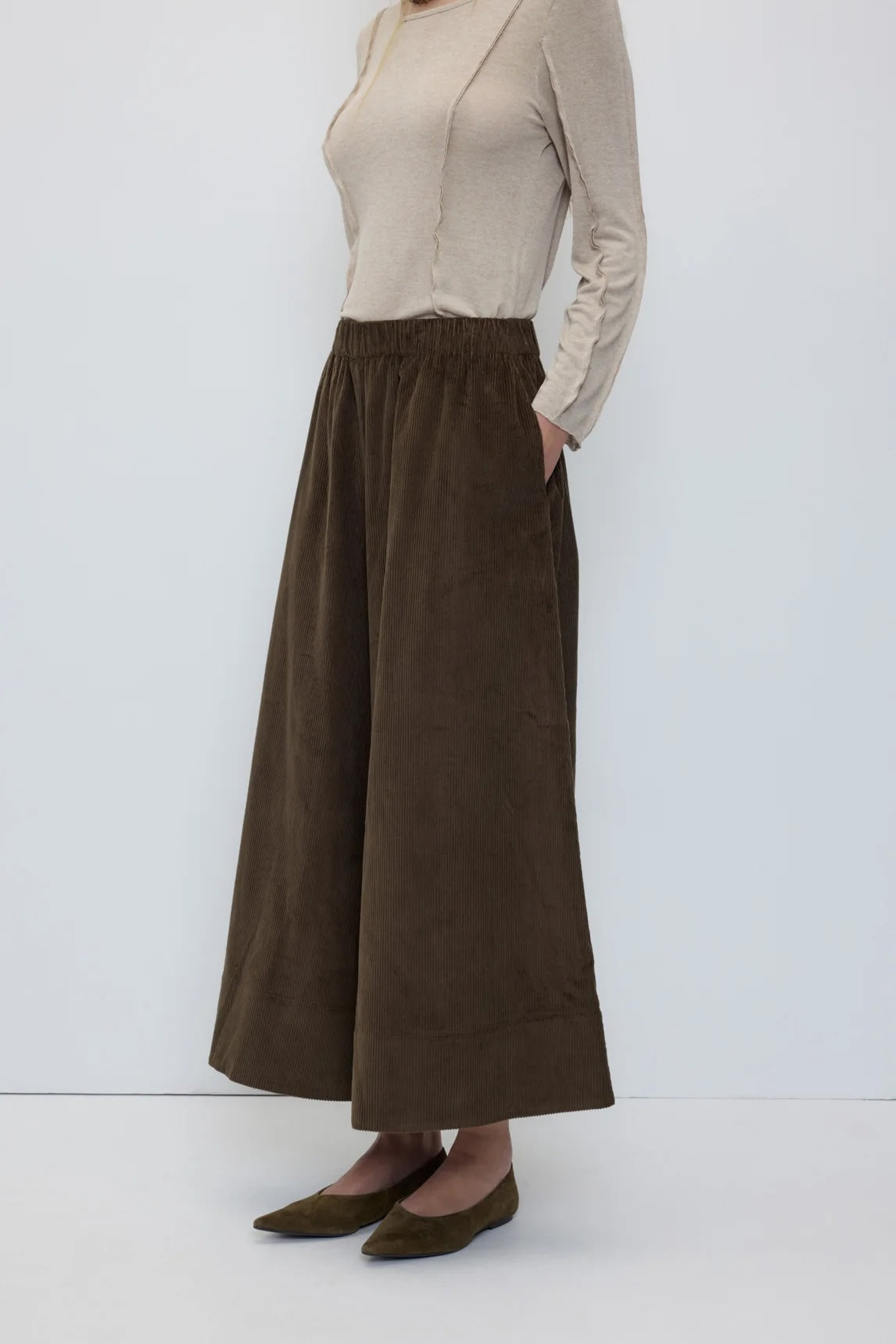 A model wearing a beige long-sleeve top and a dark brown wide-leg corduroy skirt with side pockets, paired with olive green flats, against a plain white studio background.