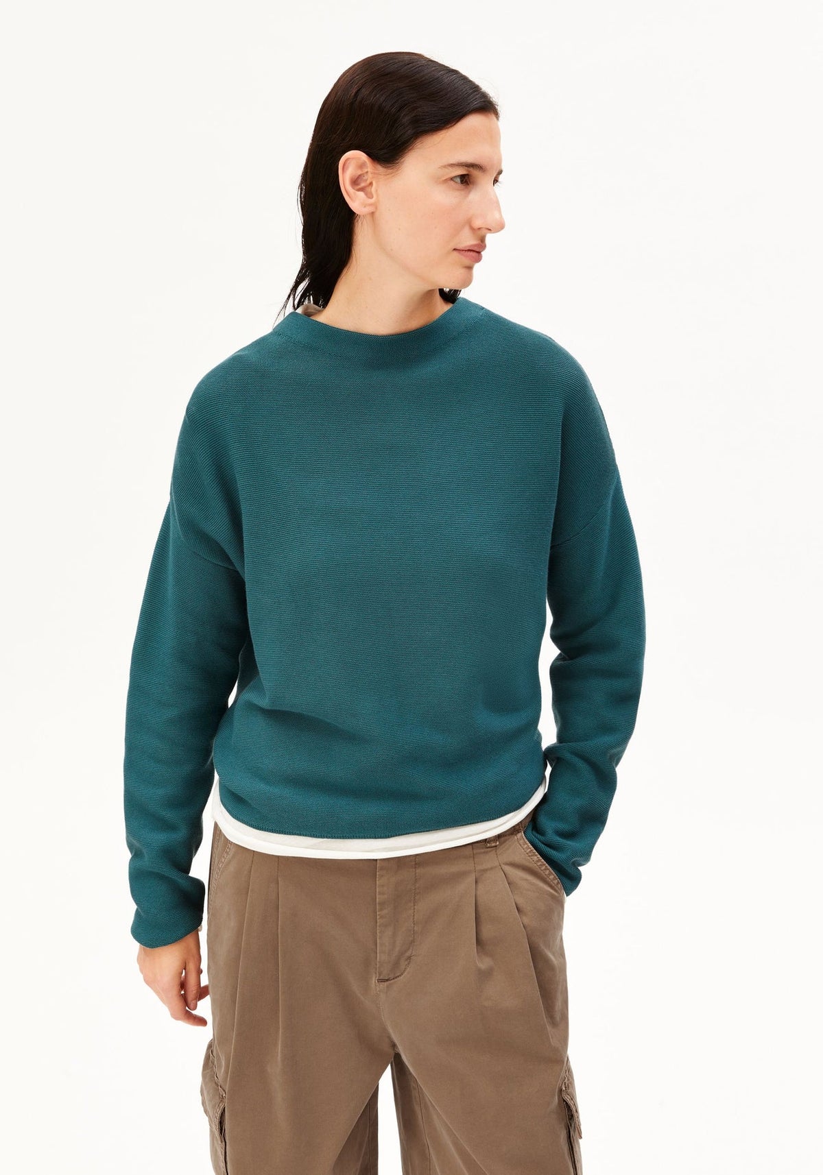A model wearing a dark teal knit crewneck sweater over a white undershirt and brown cargo pants, photographed against a clean white studio background.