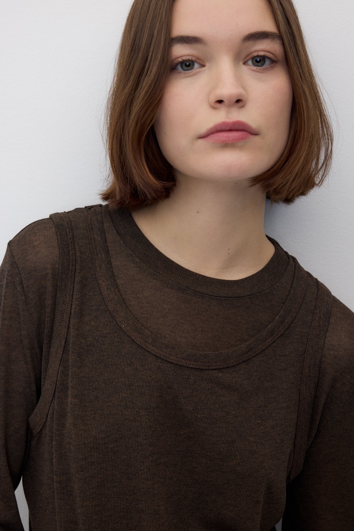 A close-up shot of a model wearing a dark brown long-sleeve crewneck top with a layered, scalloped collar detail, set against a plain light gray background.