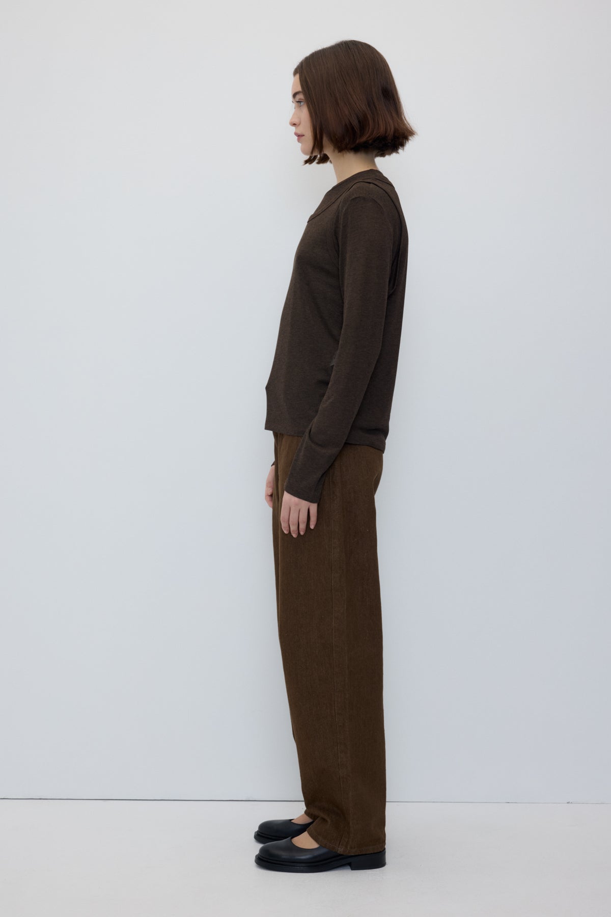 A model shown in profile wearing a dark brown long-sleeve knit top and matching wide-leg trousers, paired with black flat shoes, against a plain light gray studio background.