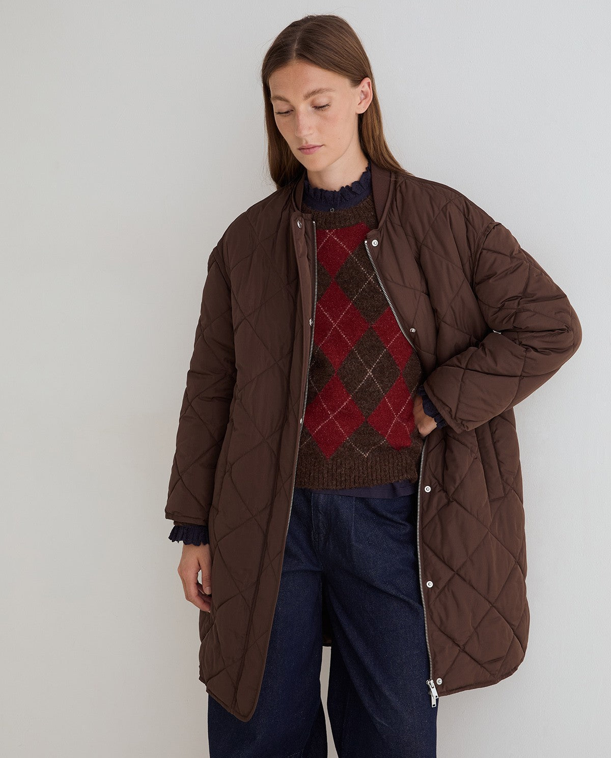 A model wearing a brown diamond-quilted puffer jacket over a red and brown argyle sweater, paired with dark blue jeans, against a plain light gray background.