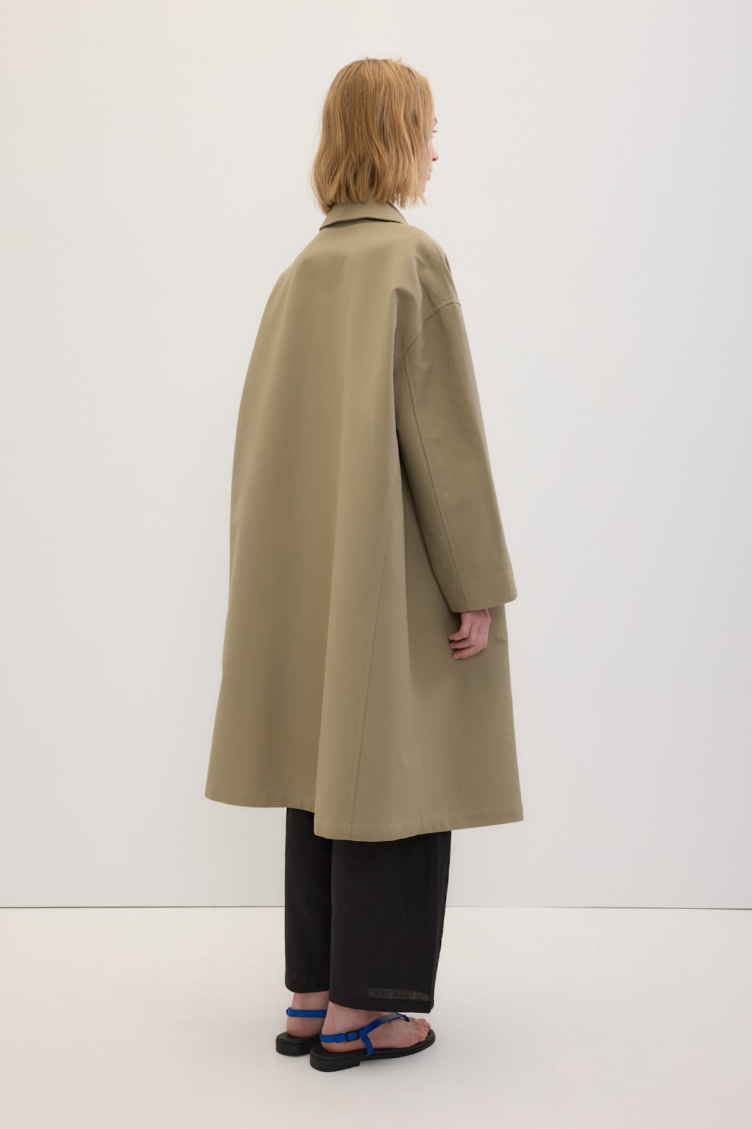 A model shown from the back wearing a long, oversized beige trench coat over black wide-leg pants and blue sandals, set against a plain white studio background.