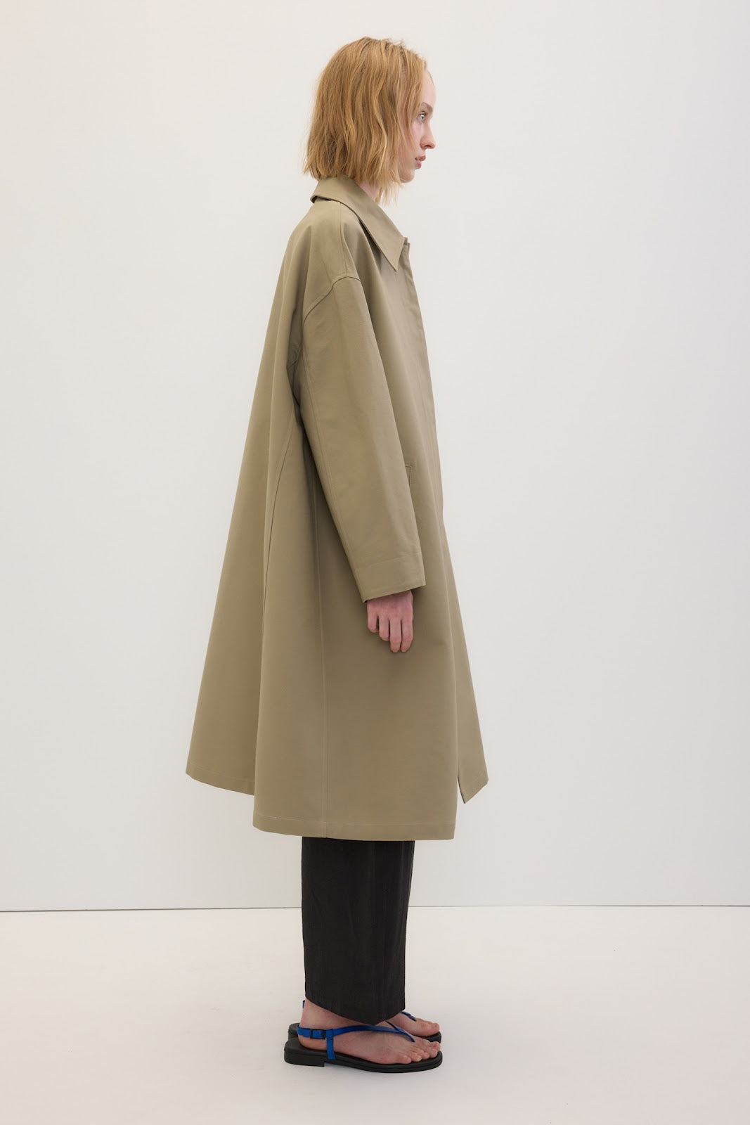 A model shown from the side profile wearing a long, oversized beige trench coat, black wide-leg pants, and blue sandals against a plain white studio background.
