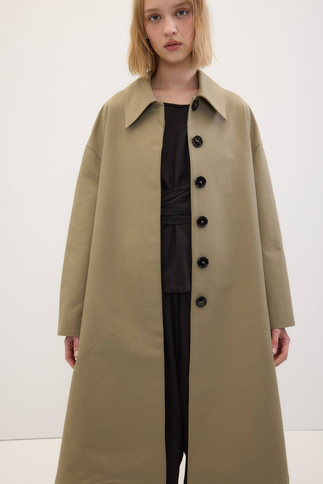 A model wearing a long, oversized beige trench coat over a black dress, photographed against a clean white studio background.