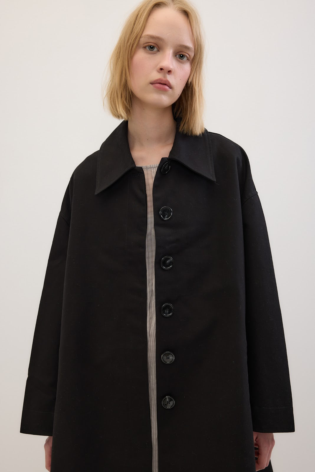 A model with short blonde hair wearing a black oversized coat over a light gray top, photographed against a plain light gray studio background.
