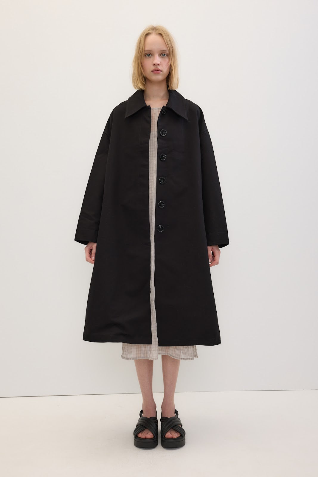 A model stands against a plain white studio background wearing a long black coat over a light dress and black sandals.