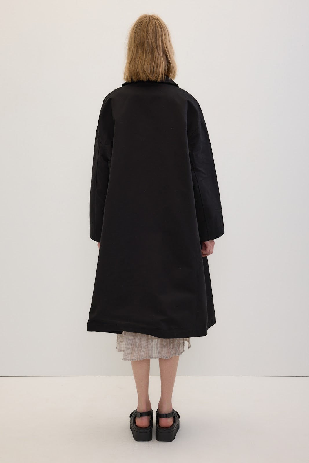 A model viewed from behind wearing a long black coat over a light-colored pleated dress and black shoes, standing against a plain white studio background.