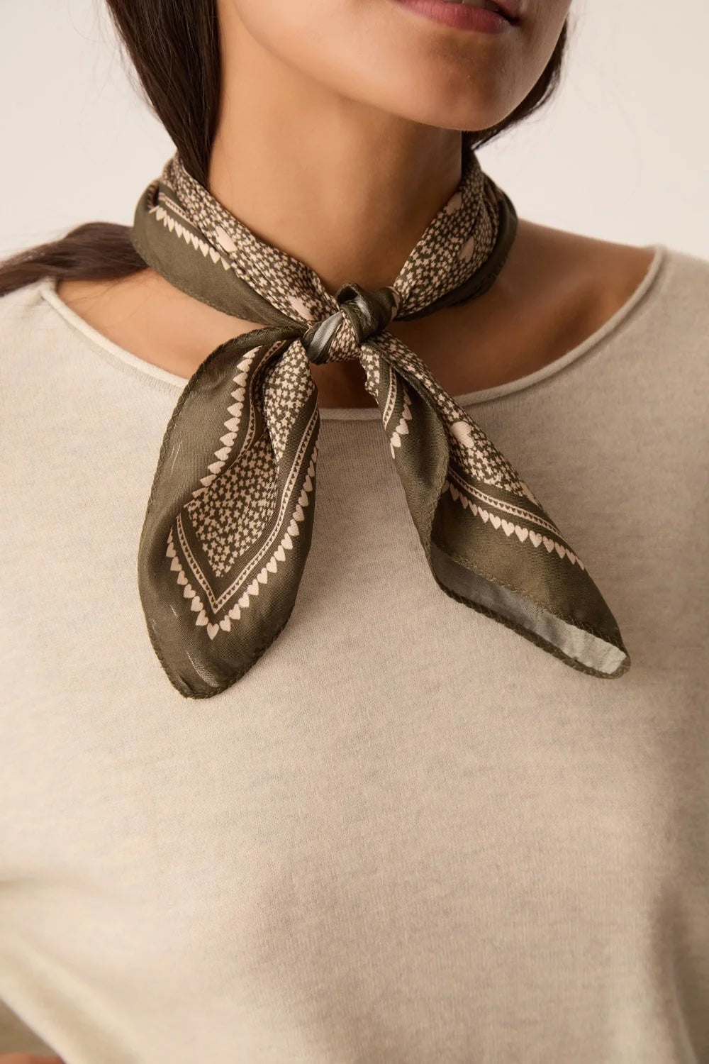 A close-up of a model wearing a dark olive green silk scarf with a beige geometric pattern, tied around her neck over a light beige top.