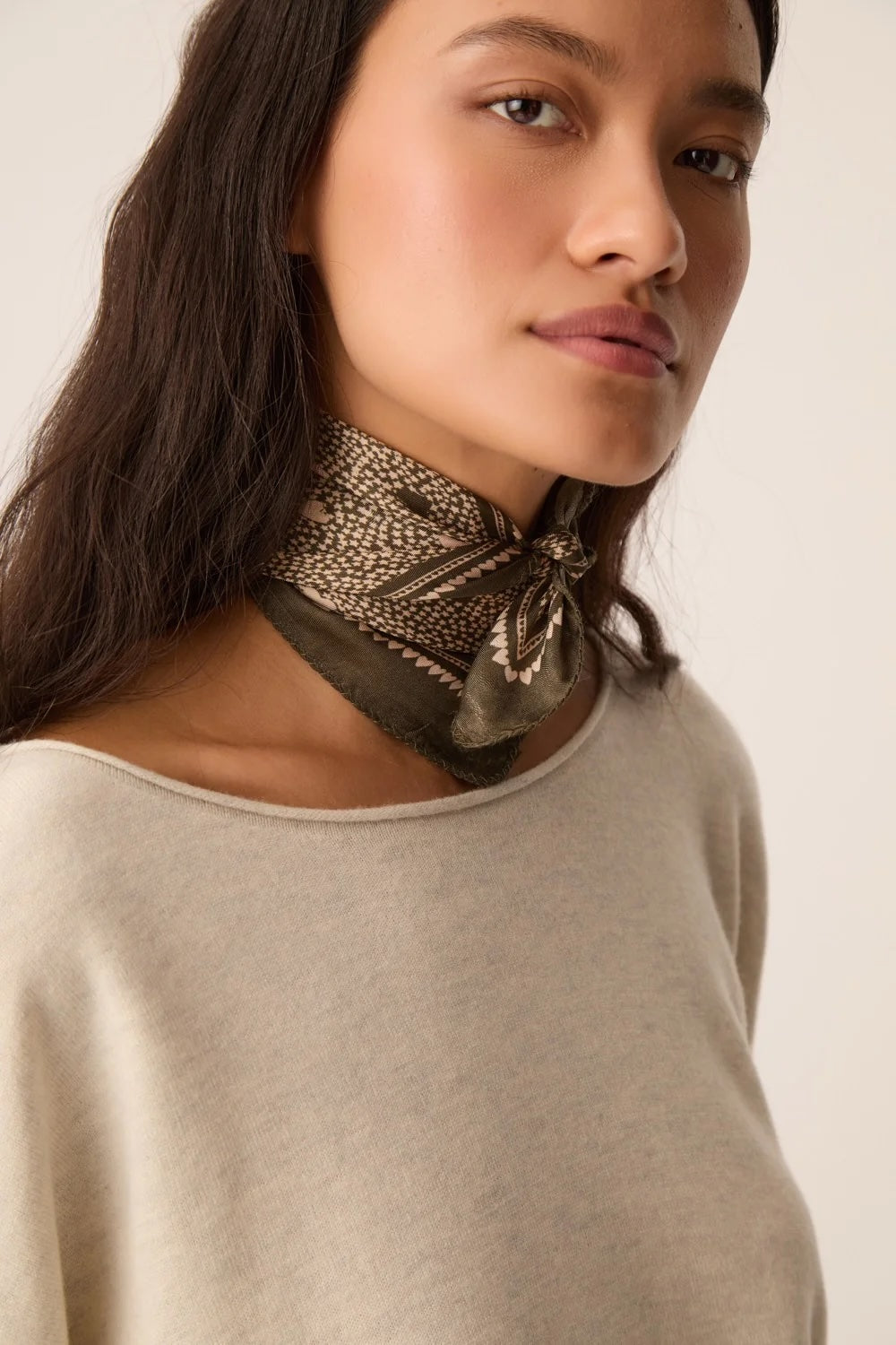 A close-up of a woman wearing a beige knit top and a patterned olive green and brown neck scarf tied at the collar.