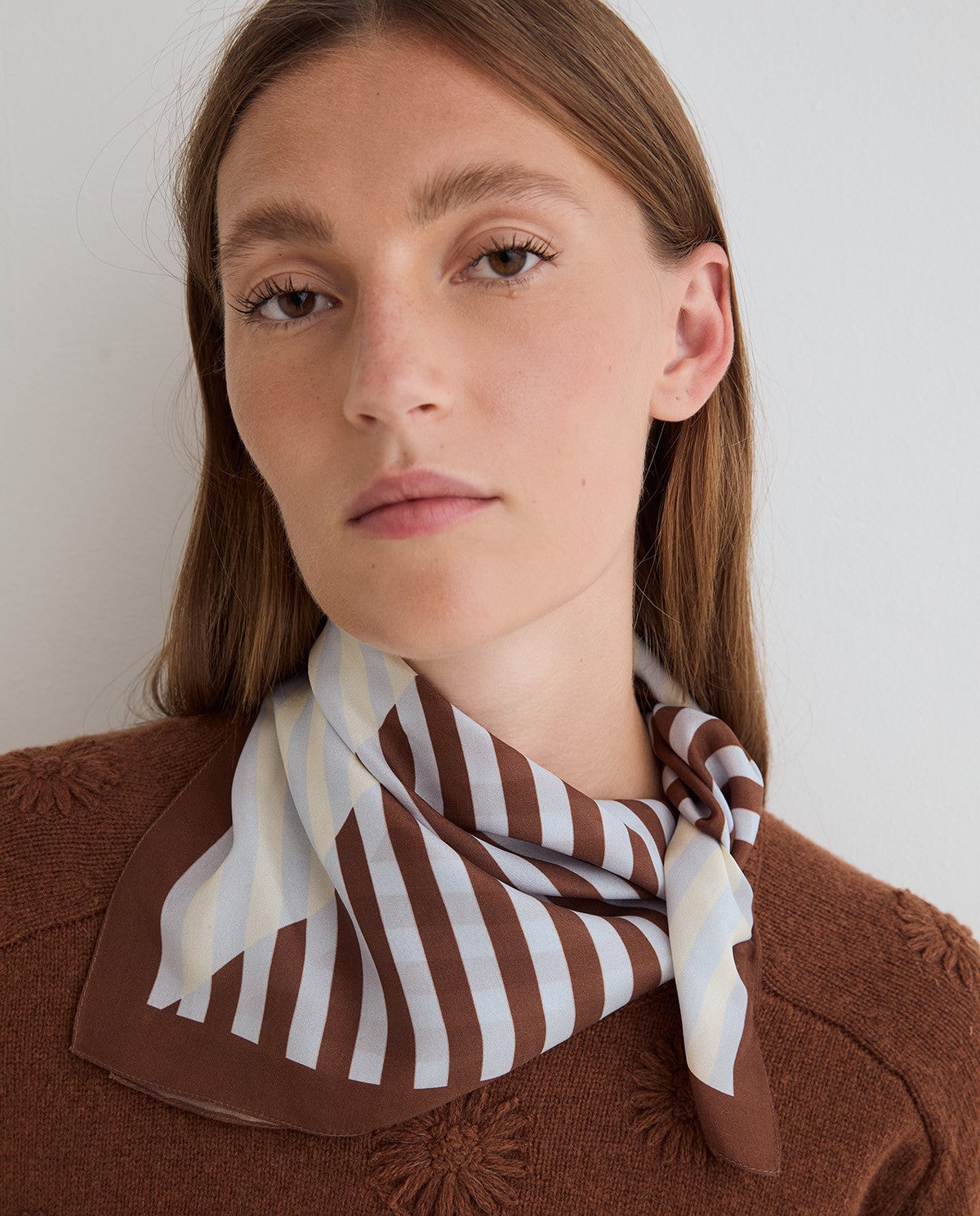 A woman wearing a brown and white striped silk scarf tied around her neck, paired with a brown textured knit sweater against a plain light gray background.