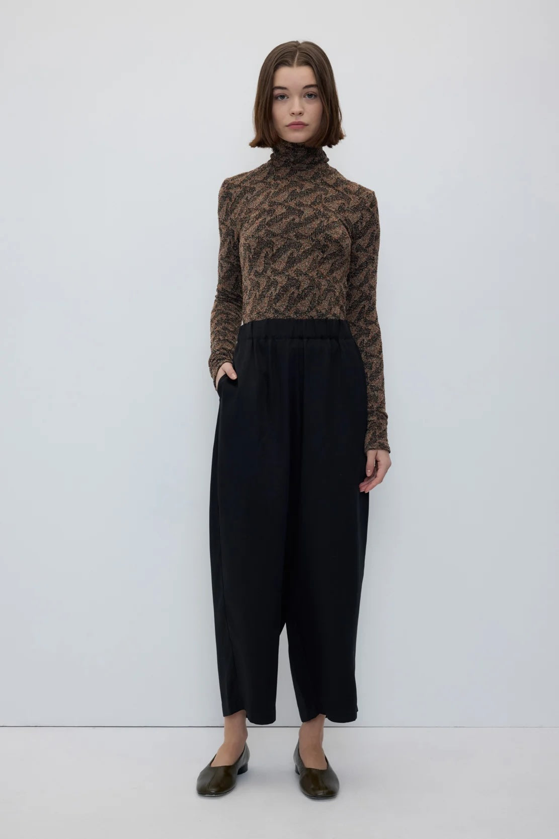 A model stands against a plain white studio background wearing a brown and black patterned mock-neck knit top, black wide-leg cropped pants, and dark brown shoes.
