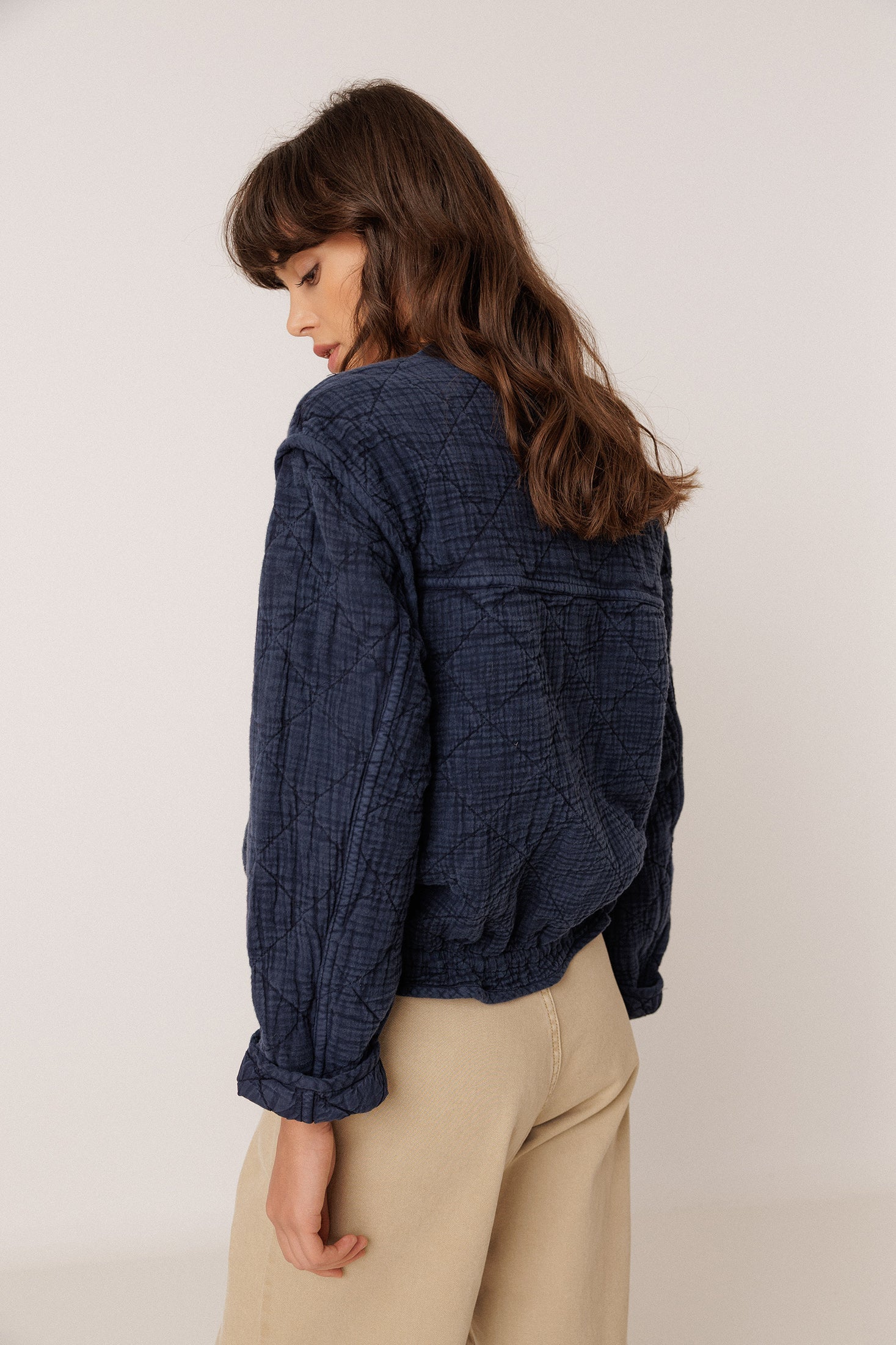 A woman wearing a dark navy textured crinkle jacket and beige trousers, photographed from a side-back angle against a plain light background.