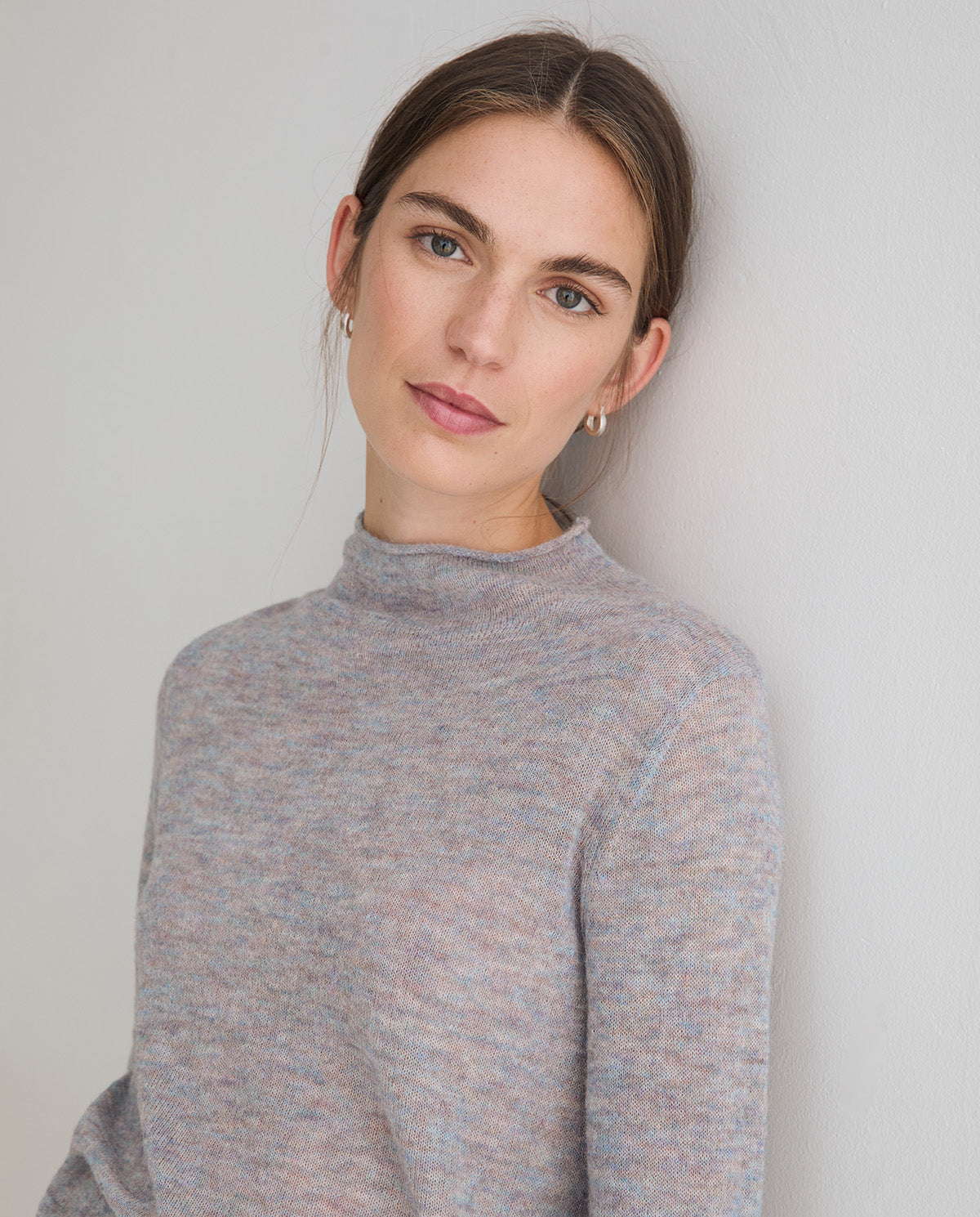 A woman with brown hair pulled back wears a light gray marled mock neck sweater against a plain light gray wall.