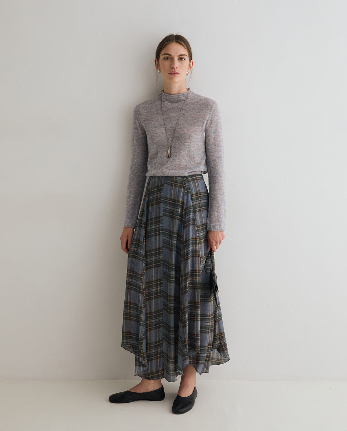 A woman stands against a plain light gray wall wearing a light gray mock-neck knit top and a long gray plaid skirt with black flats.