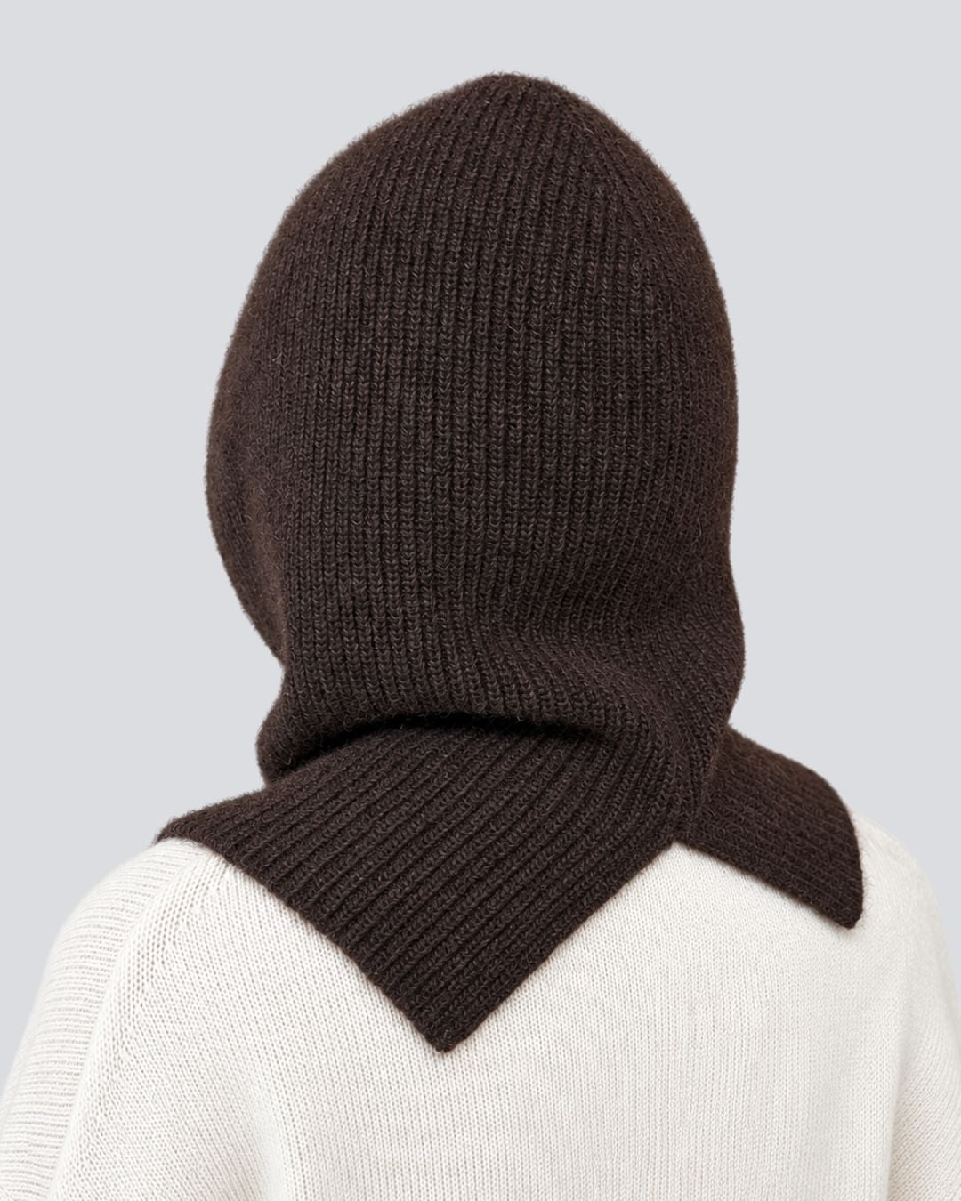A dark brown ribbed knit hooded balaclava or neck warmer shown on a model from the back, paired with a cream-colored knit sweater against a light gray background.