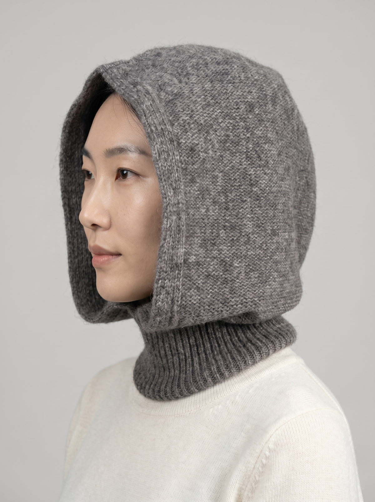 A woman wearing a gray knit hooded neck warmer with a ribbed turtleneck collar, shown in profile against a light gray background.
